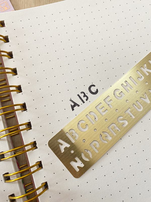 Caderno com folhas pontilhadas e régua de letras dourada com a palavra ABC carimbada