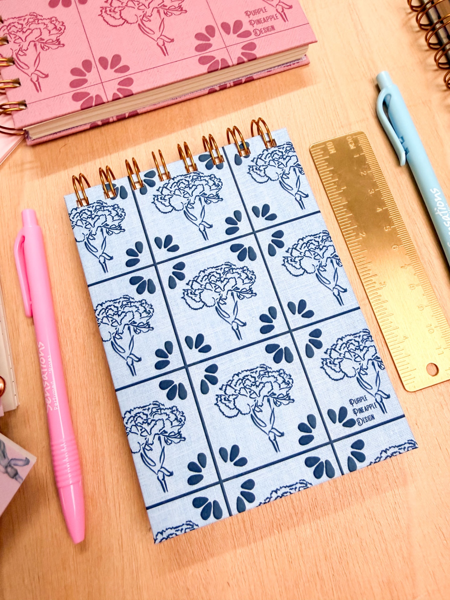 Caderno espiral com capa azul clara decorada com padrão floral e texto PURPLE PINEAPPLE DESIGN