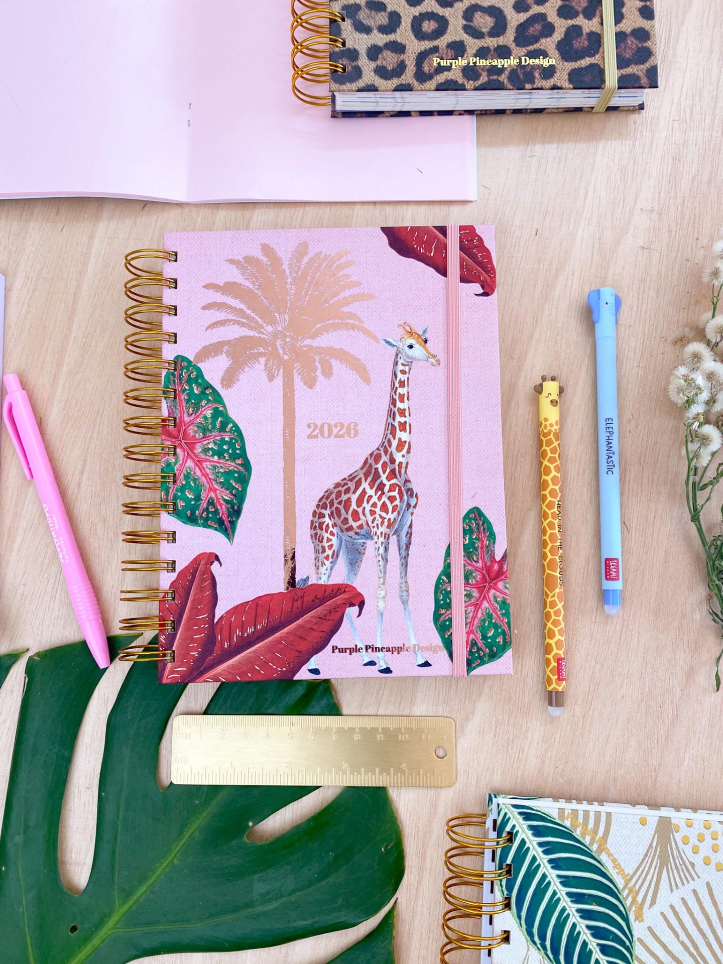Caderno 2023 estilo tropical com girafa e folhas, canetas e lápis decorativos, régua dourada, sobre mesa de madeira