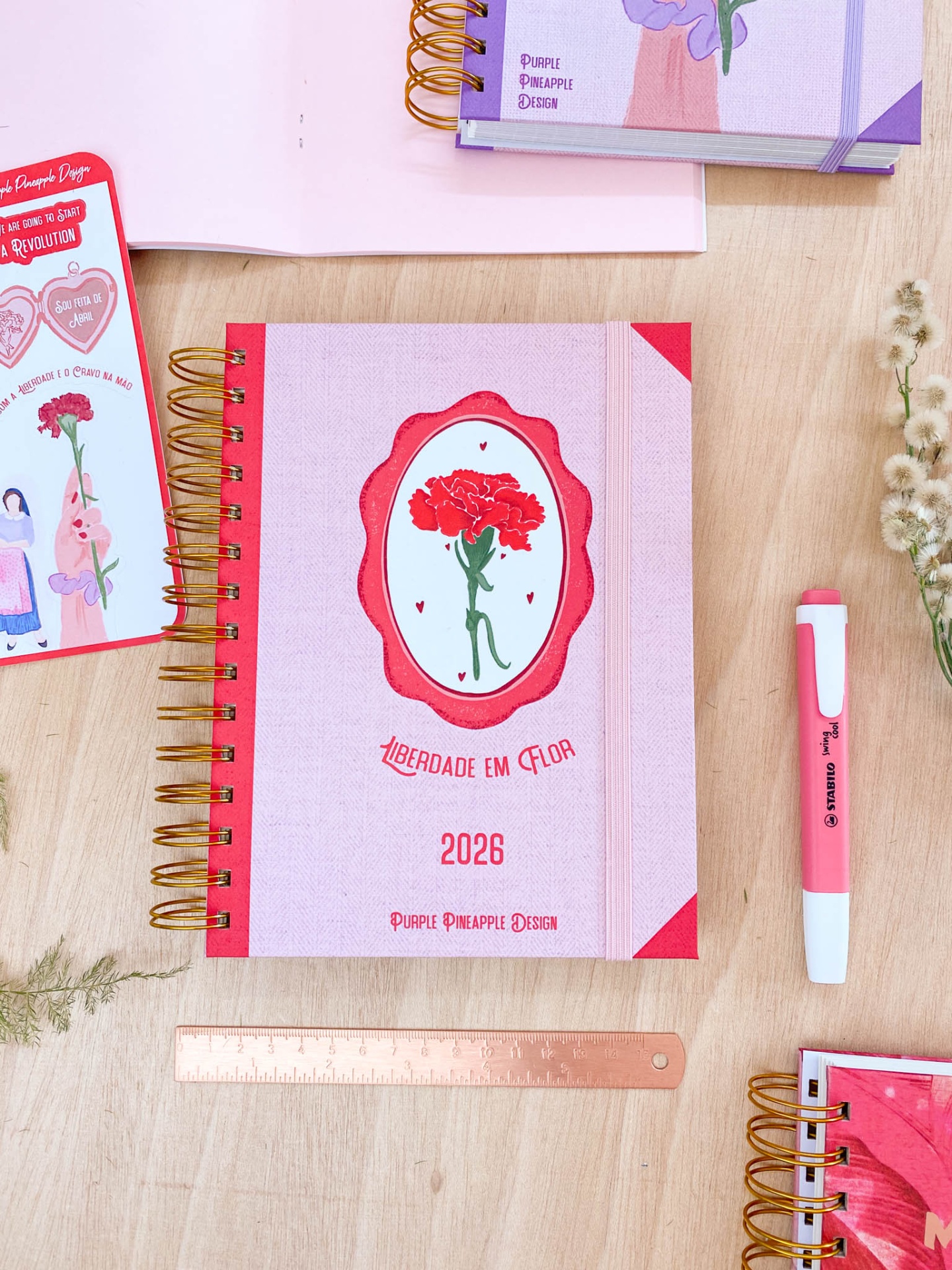Caderno espiral rosa com ilustração de cravo e texto, régua, caneta e outros cadernos sobre mesa de madeira.