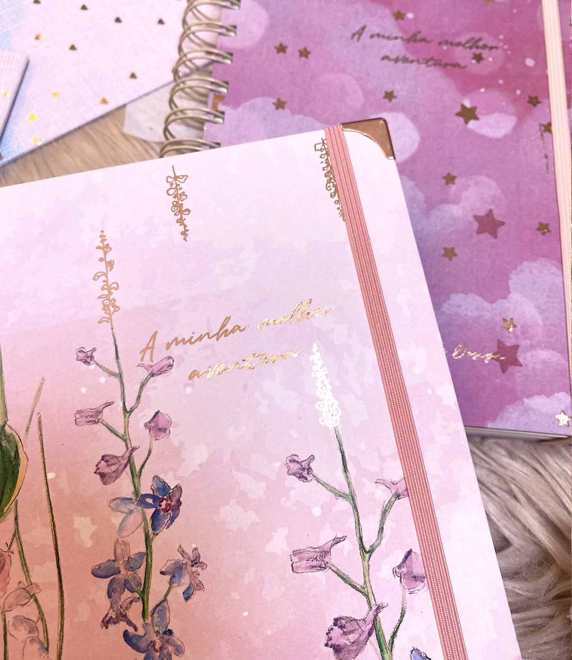Cadernos rosa e lilás com ilustrações florais e texto dourado, num tapete cinza