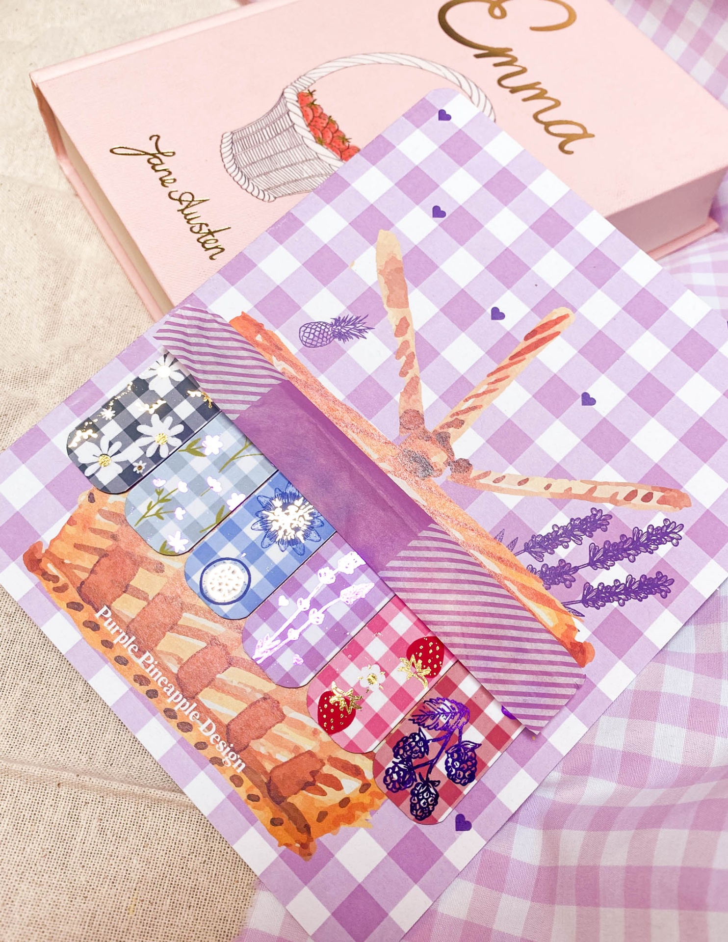 Cartão ilustrado com pães e frascos decorativos com padrão xadrez lilás, ao lado de caixa rosa com texto Emma Jane Austen