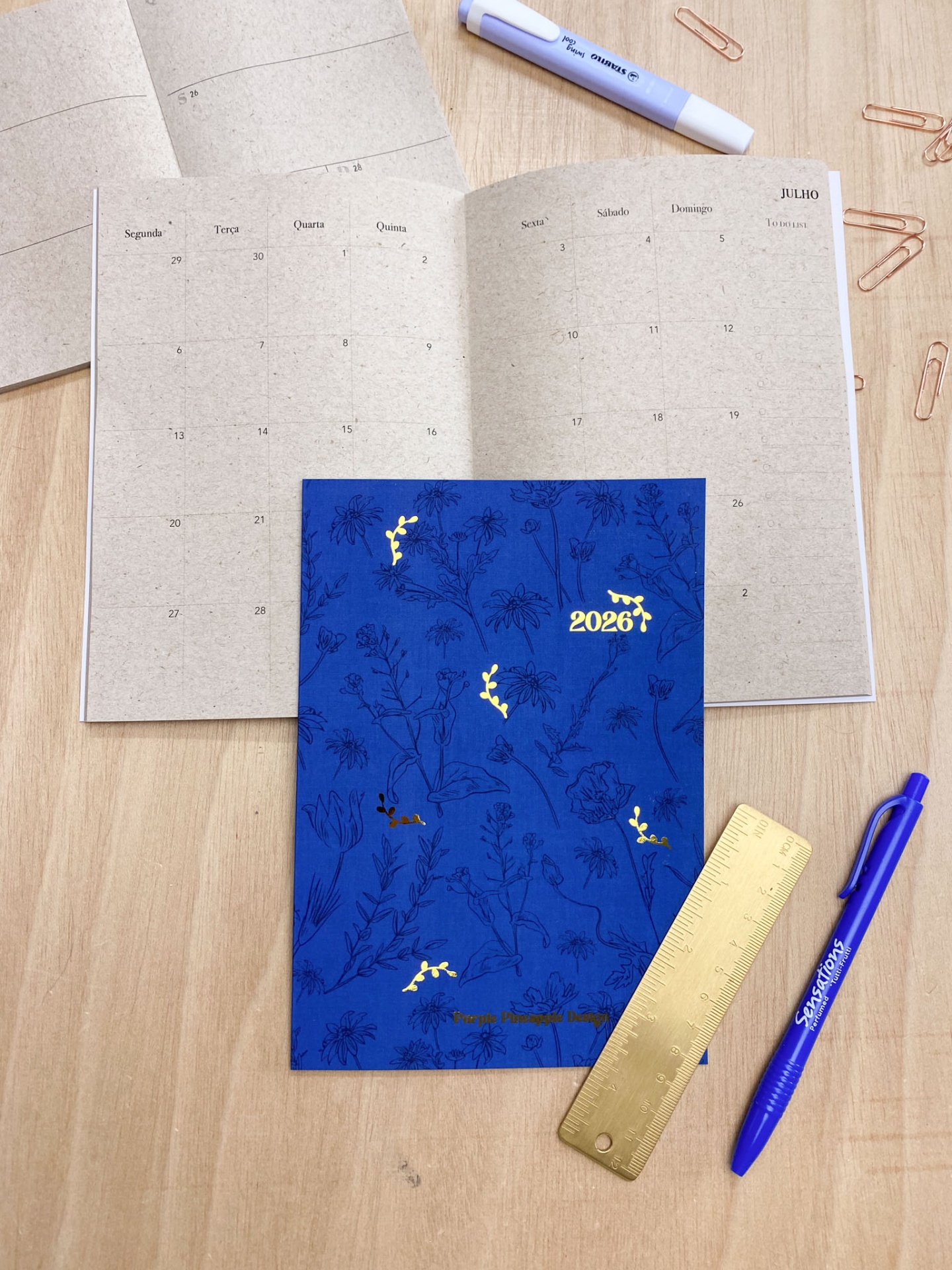 Agenda azul 2026 com ilustrações de plantas e detalhes dourados sobre mesa com régua, caneta e marcador.