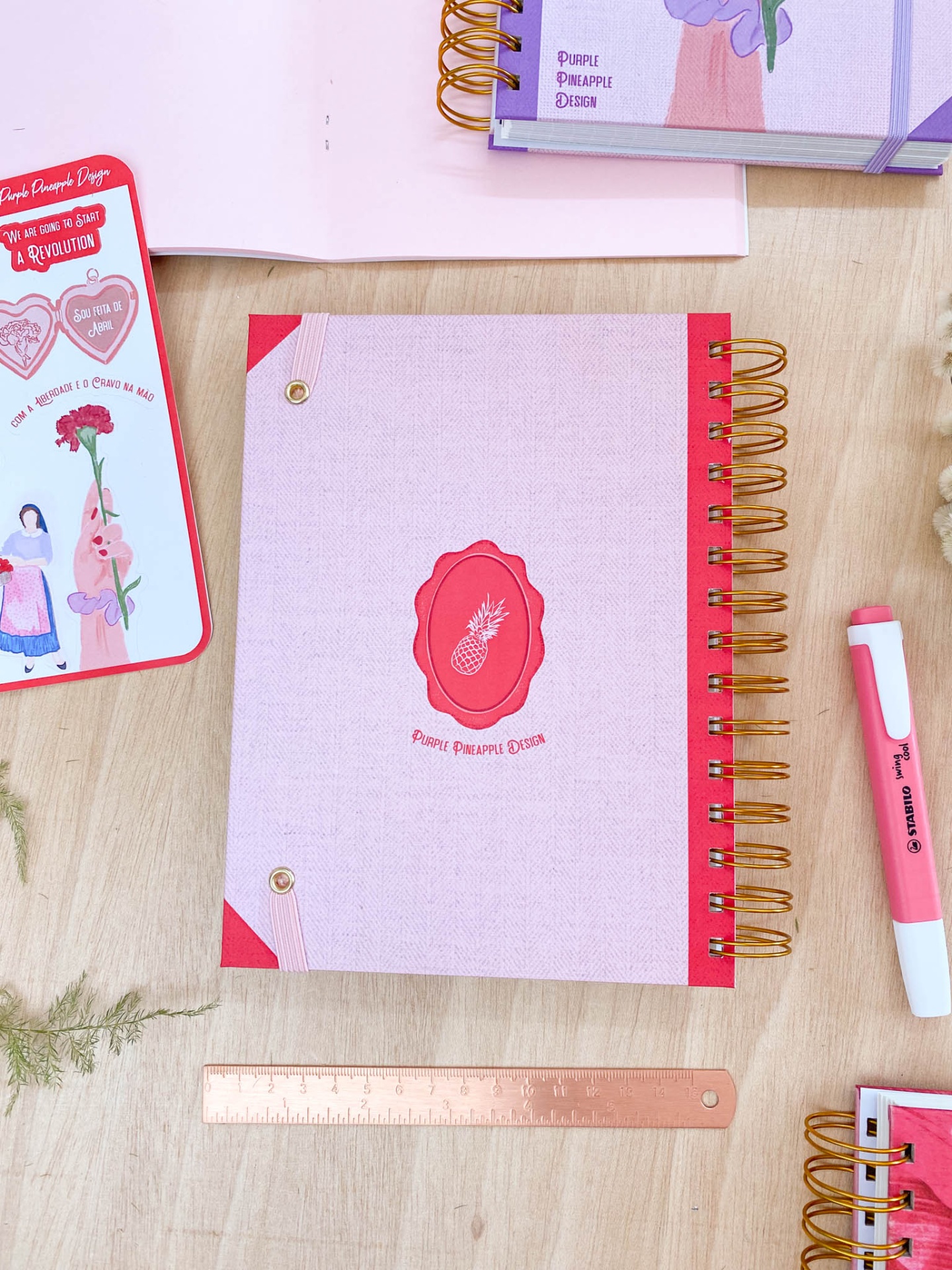 Caderno rosa com espiral dourada e selo vermelho com ananás, sobre mesa com régua e marcador