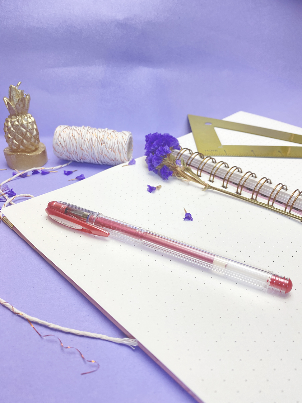Caderno aberto com caneta vermelha, régua triangular dourada, flor seca roxa e abacaxi decorativo dourado sobre fundo lilás