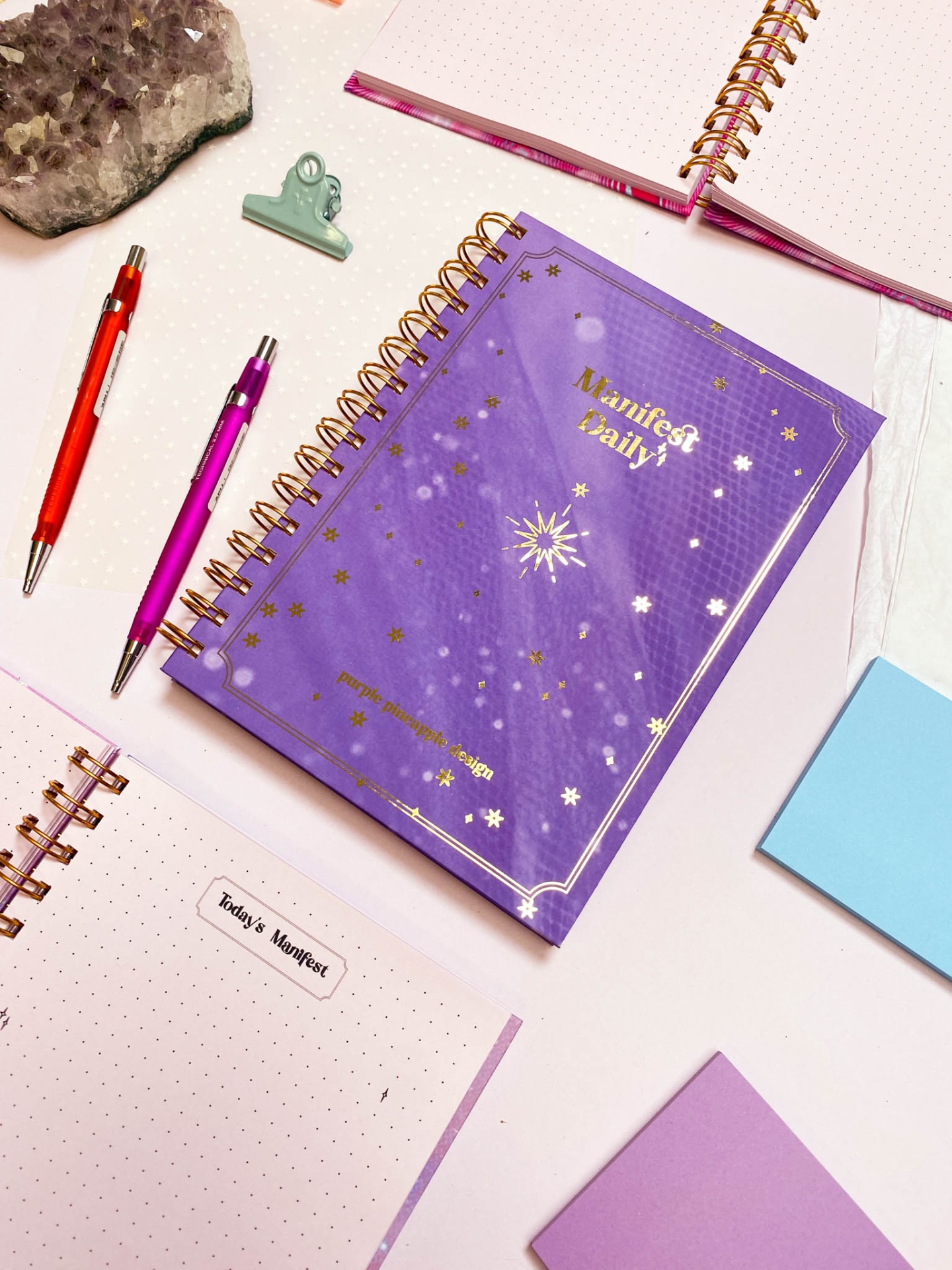Caderno roxo com capa decorada e canetas, clips, blocos de notas e pedra de ametista ao redor