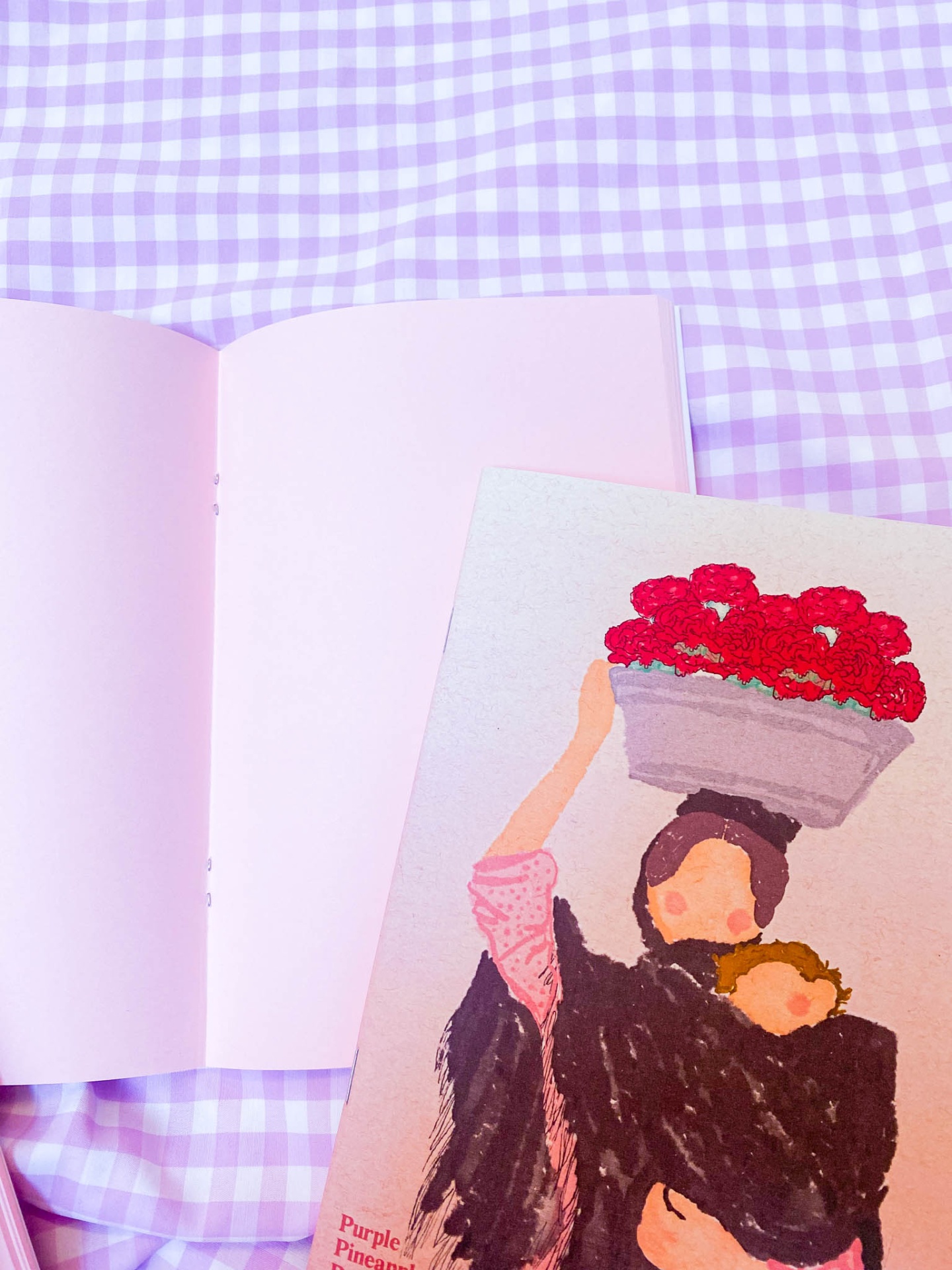 Caderno rosa aberto sobre tecido xadrez lilás e branco com ilustração colorida ao lado