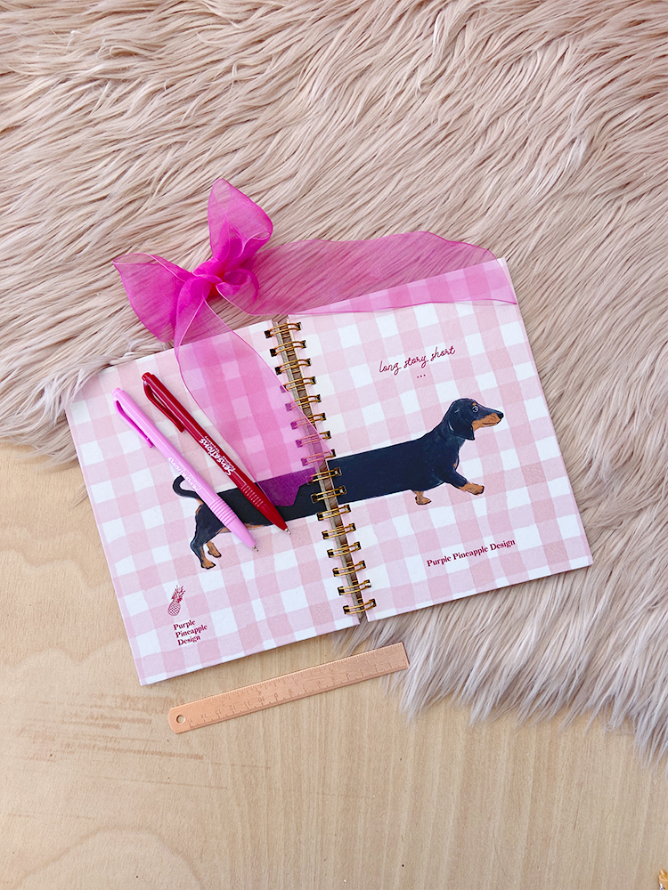 Caderno espiral com padrão xadrez rosa, cão dachshund desenhado e acessórios rosa sobre mesa