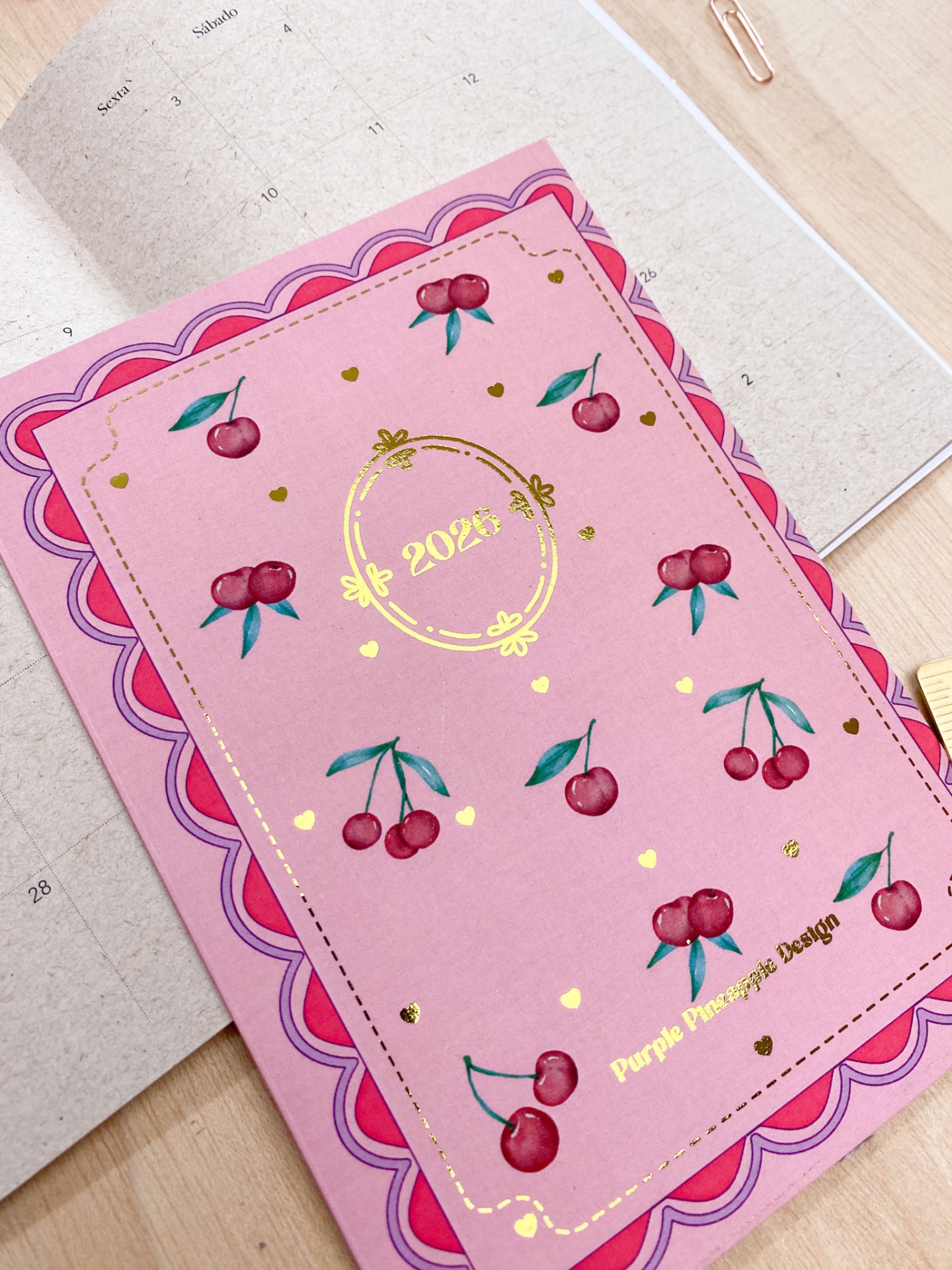 Agenda rosa com cerejas vermelhas e detalhes dourados sobre calendário