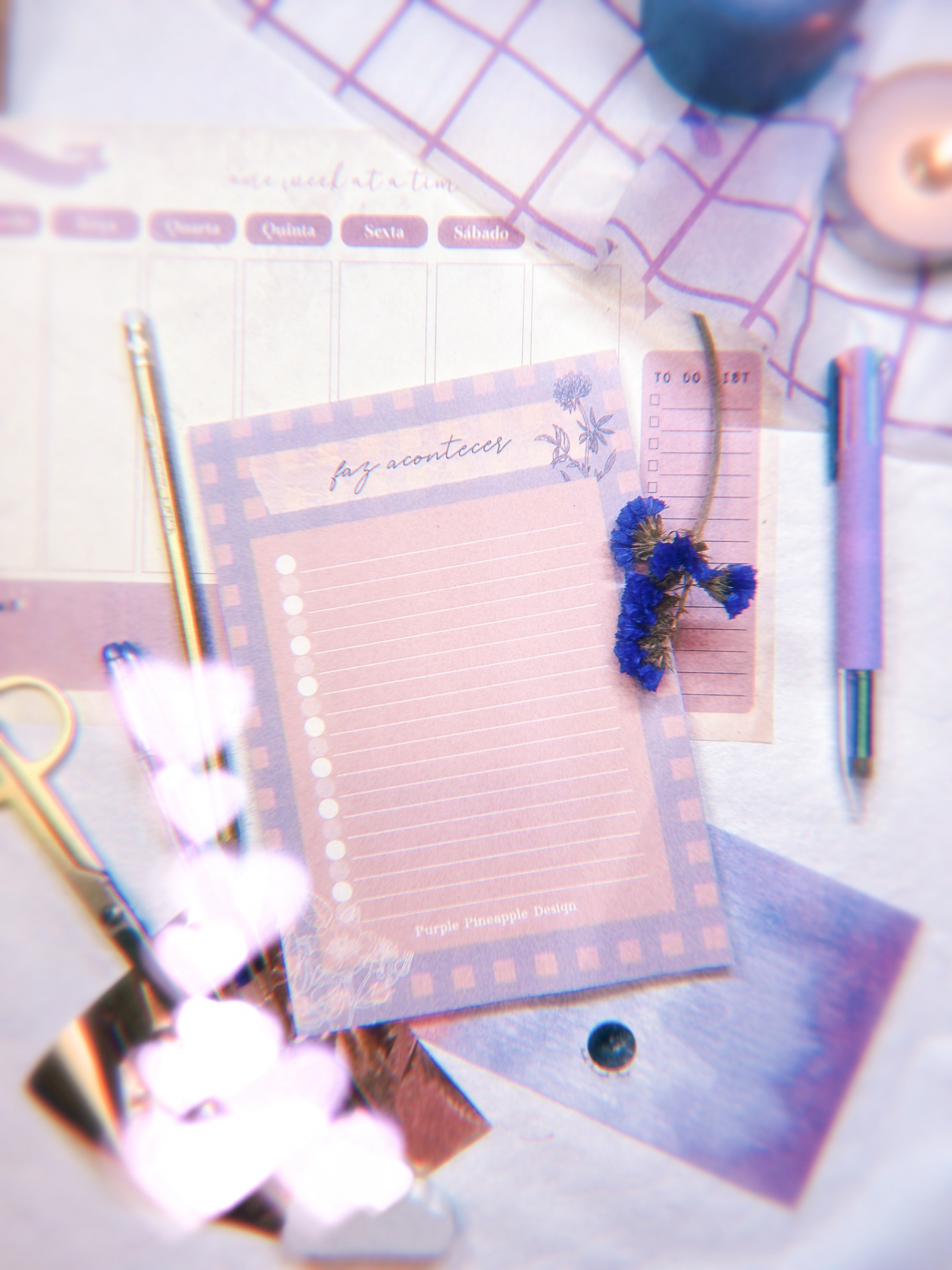Artigos de papelaria lilás e dourados com textos em português sobre superfície quadriculada.