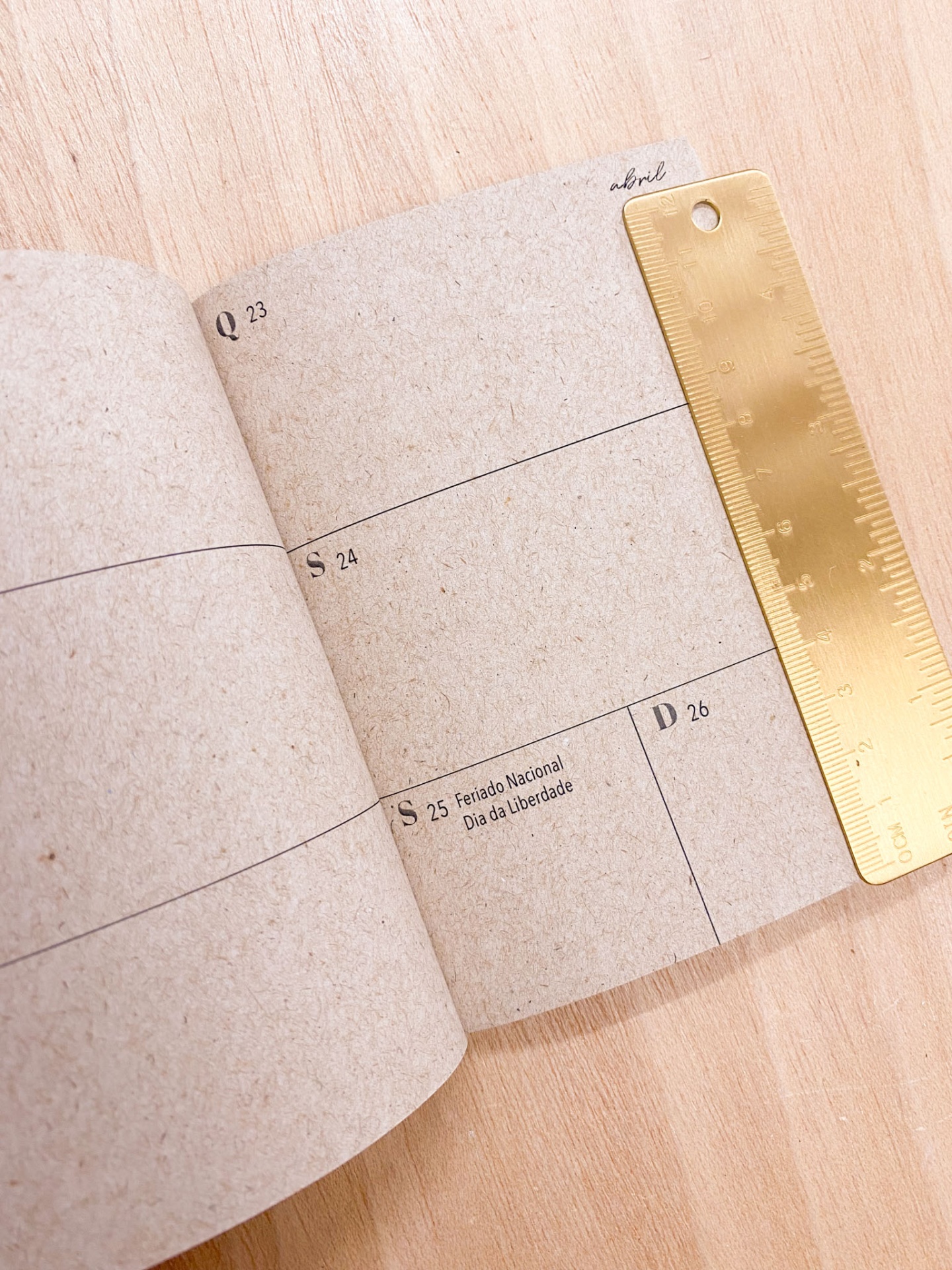 Calendário em papel reciclado com régua metálica dourada sobre mesa de madeira clara