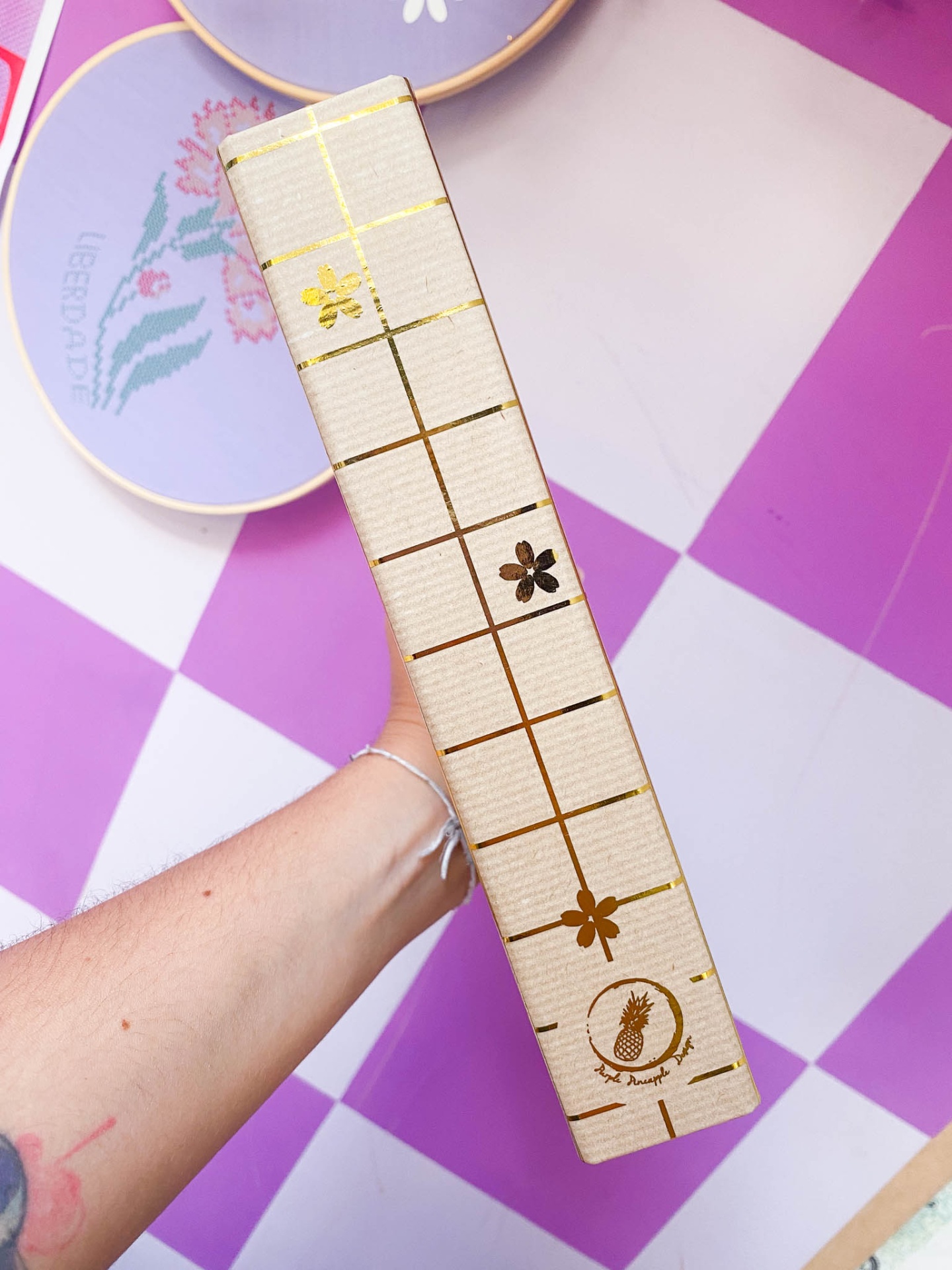 Caixa retangular bege com padrão quadriculado dourado e flores douradas segurada por uma mão sobre pano roxo e branco