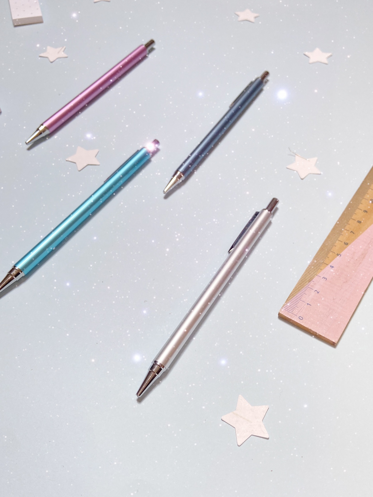Quatro canetas metálicas coloridas e duas réguas de madeira sobre superfície cinza com estrelas brancas