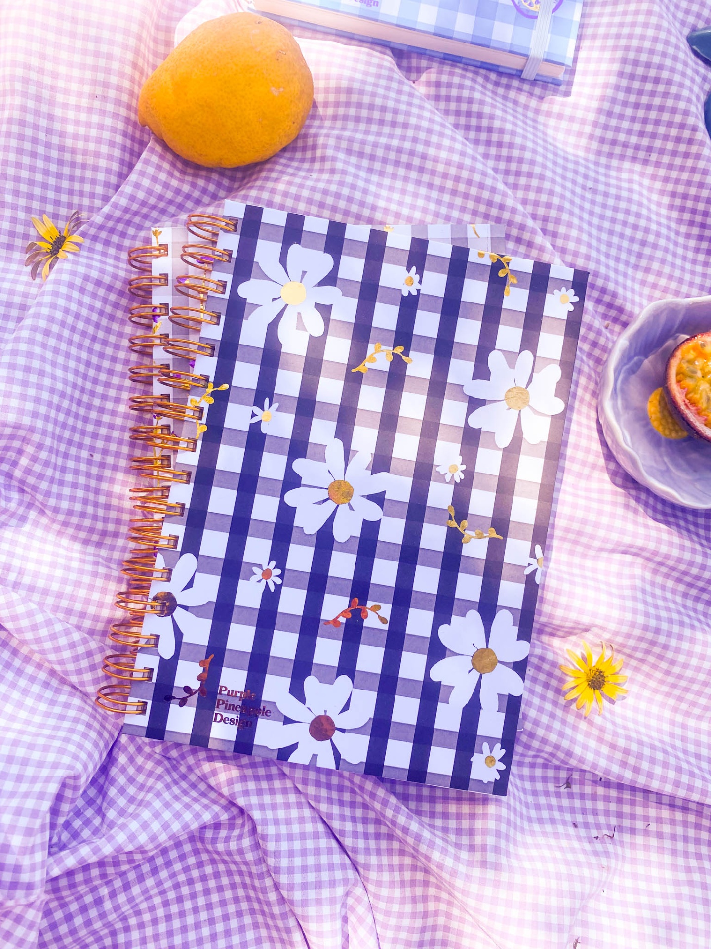 Dois cadernos espiralados com estampado xadrez azul e branco e flores, sobre tecido lilás xadrez.