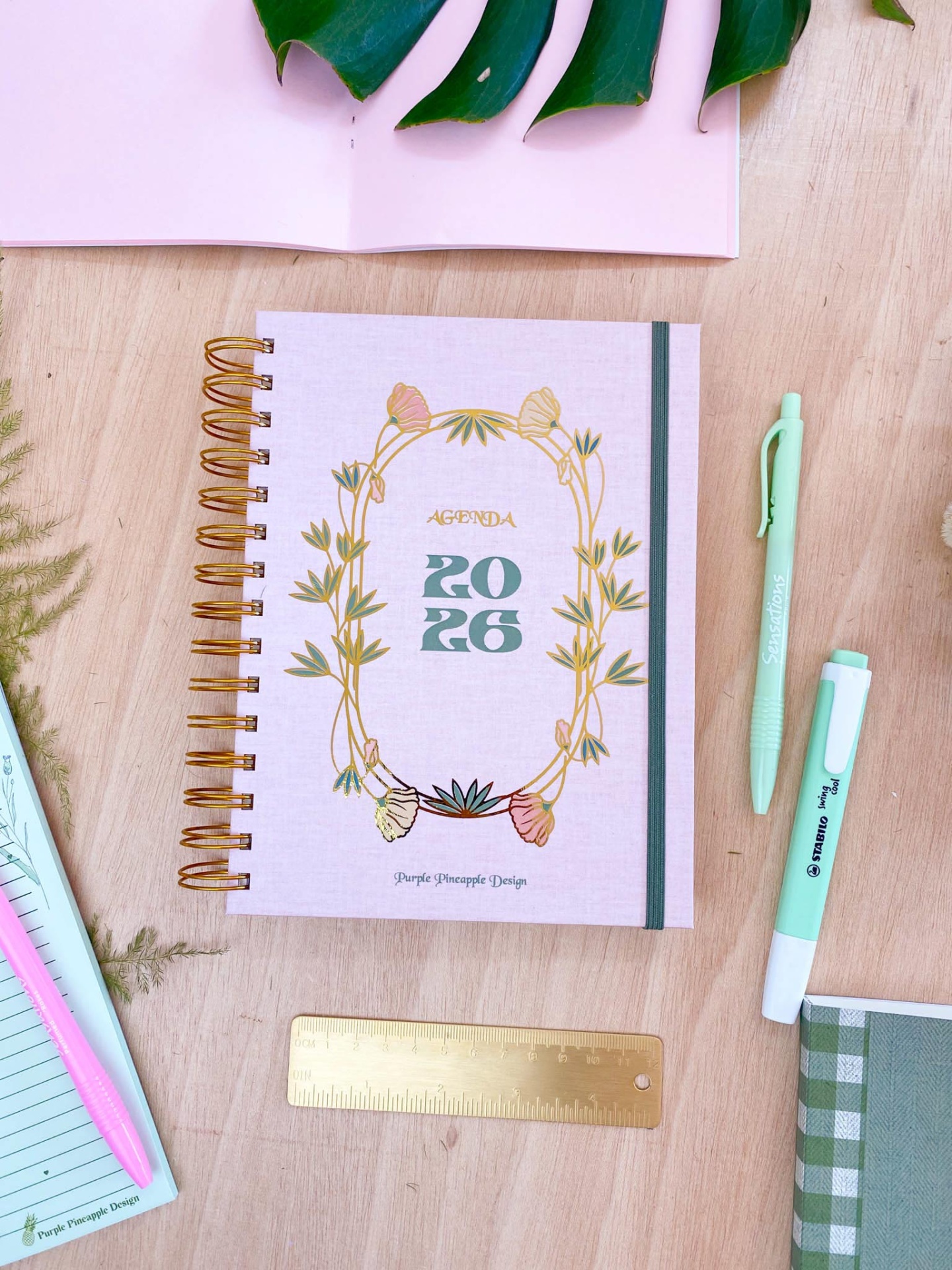 Agenda 2026 com capa rosa claro e decoração floral, canetas e régua em superfície de madeira clara.
