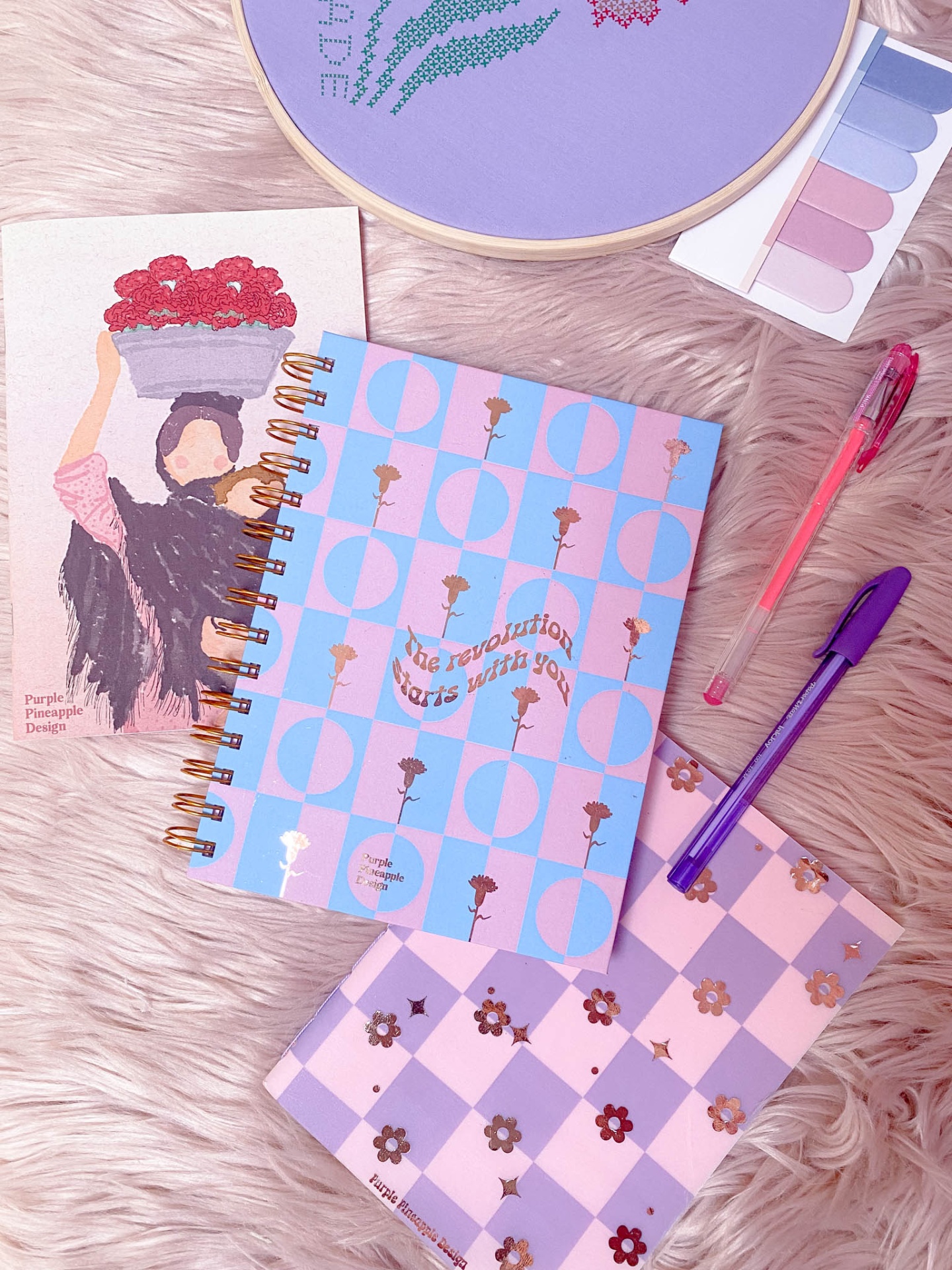 Cadernos decorados, canetas, paleta de sombras e cartão ilustrado sobre superfície peluda rosa.