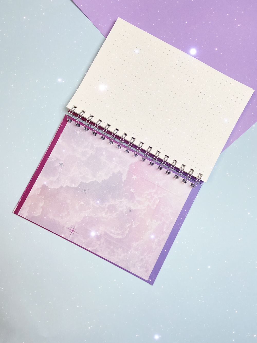 Caderno espiral com capa decorada e páginas internas em branco sobre fundo lilás e azul.