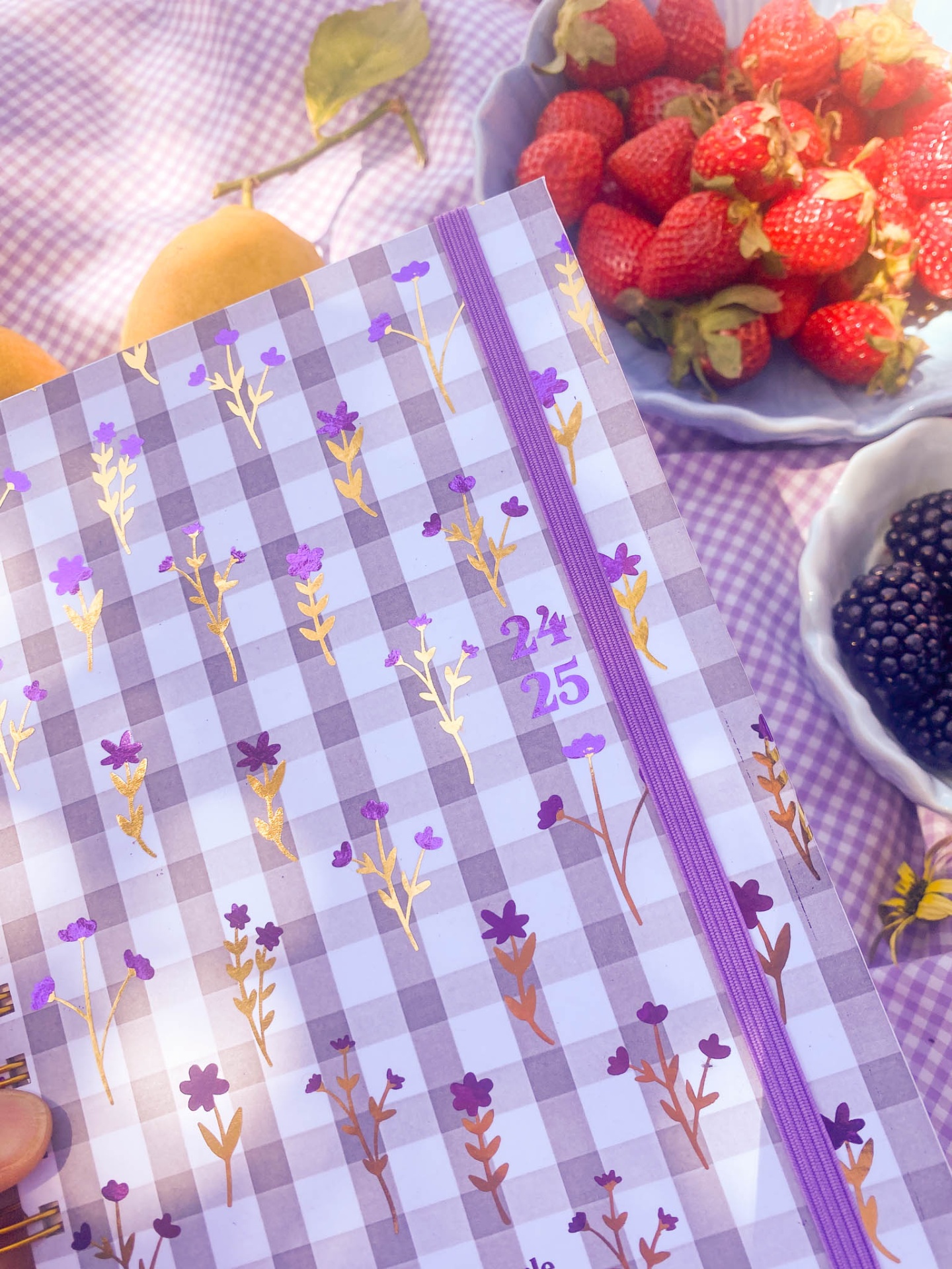 Agenda floral lilás sobre toalha xadrez com frutas em tigelas ao fundo