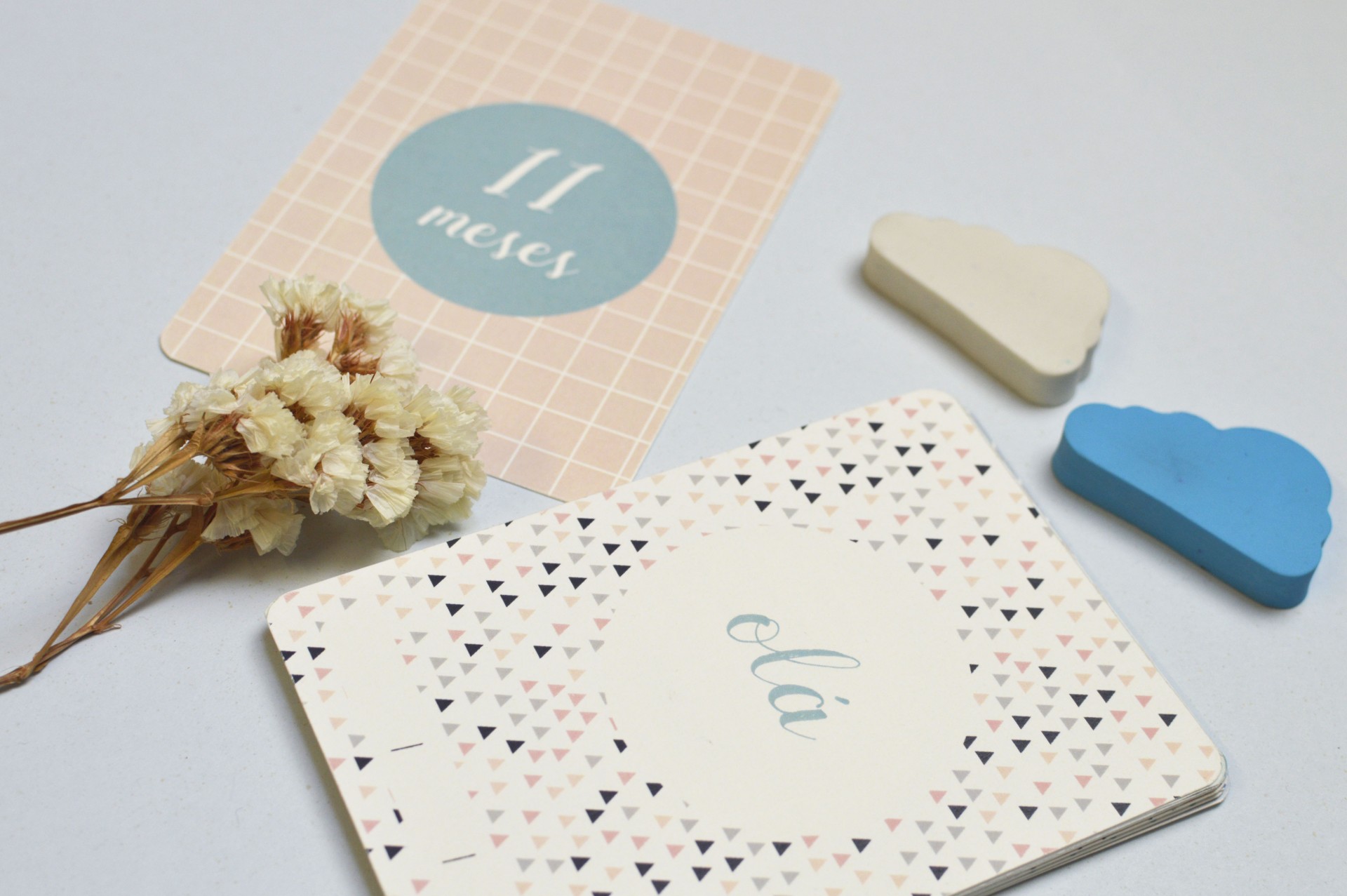 Cartões de marco de bebé com texto em português, flores secas e objetos em forma de nuvem sobre superfície branca.