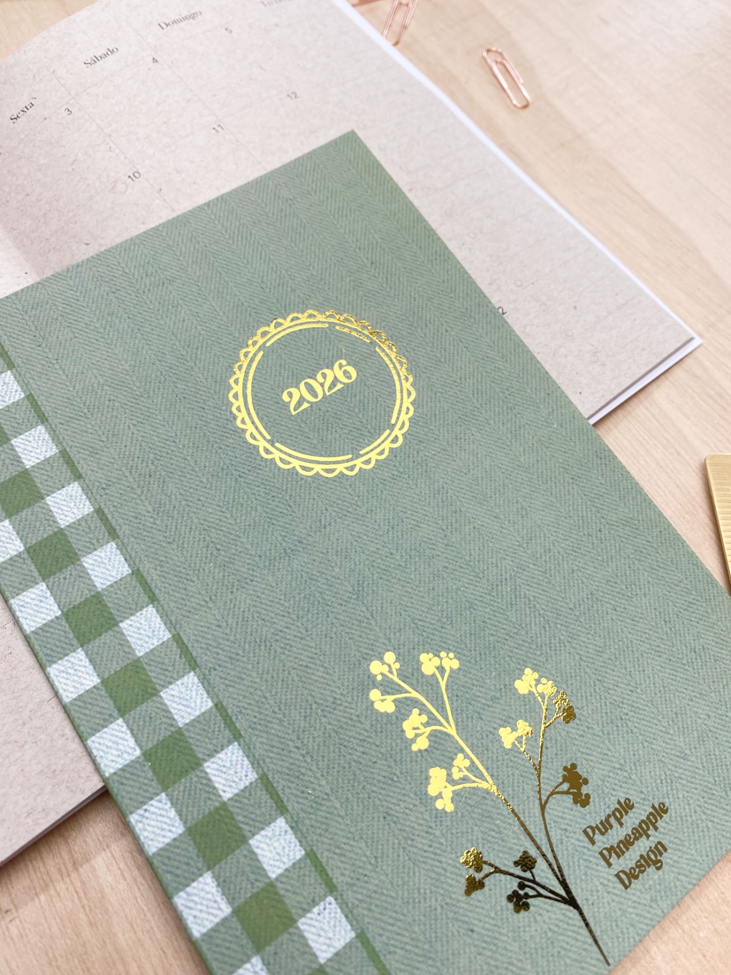 Agenda verde com padrão xadrez e detalhes dourados sobre mesa de madeira clara