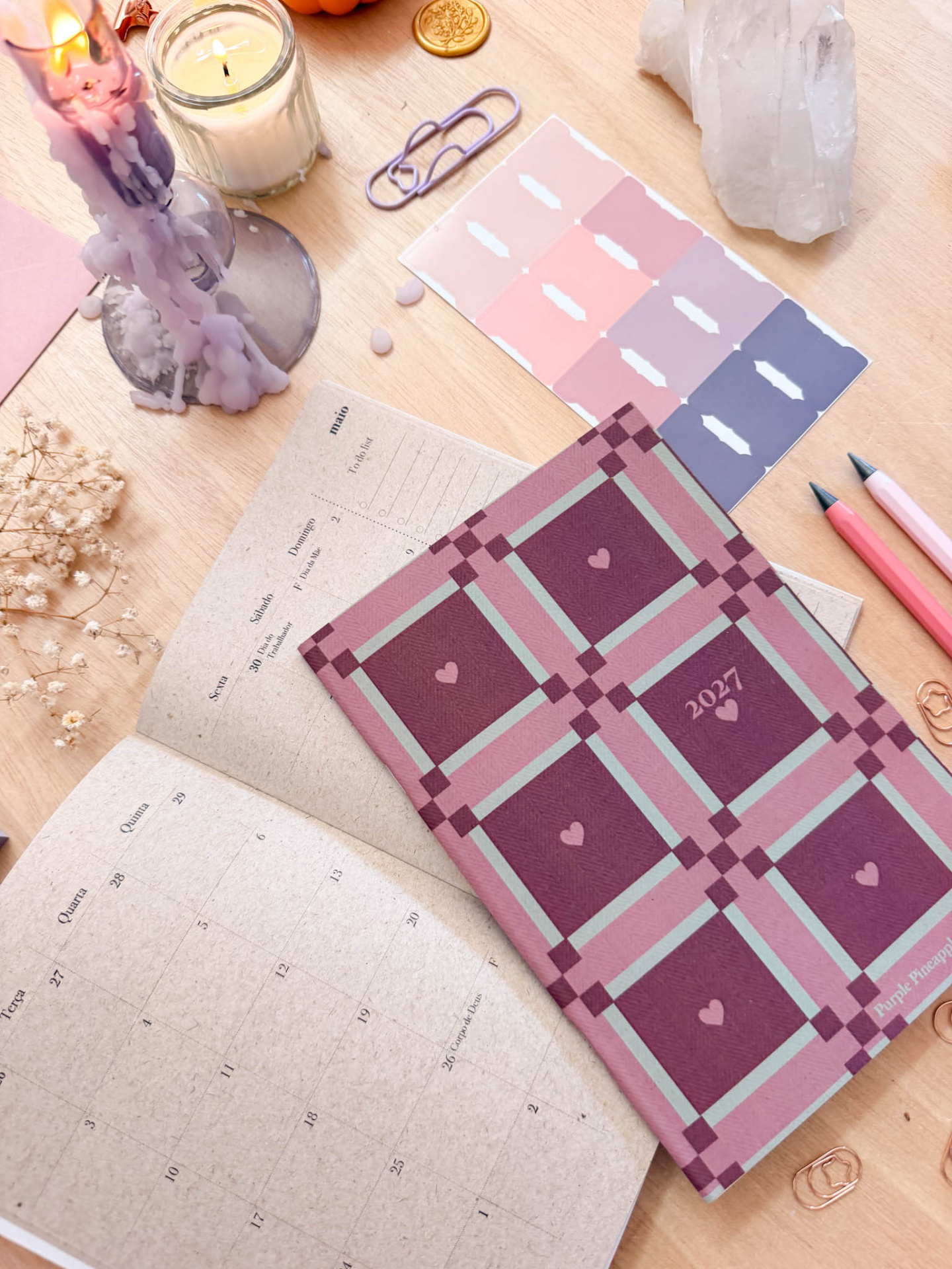 Agenda 2027 padrão roxo e rosa, calendário aberto, clips, lápis, vela acesa, pedra cristal e etiquetas adesivas em mesa de madeira