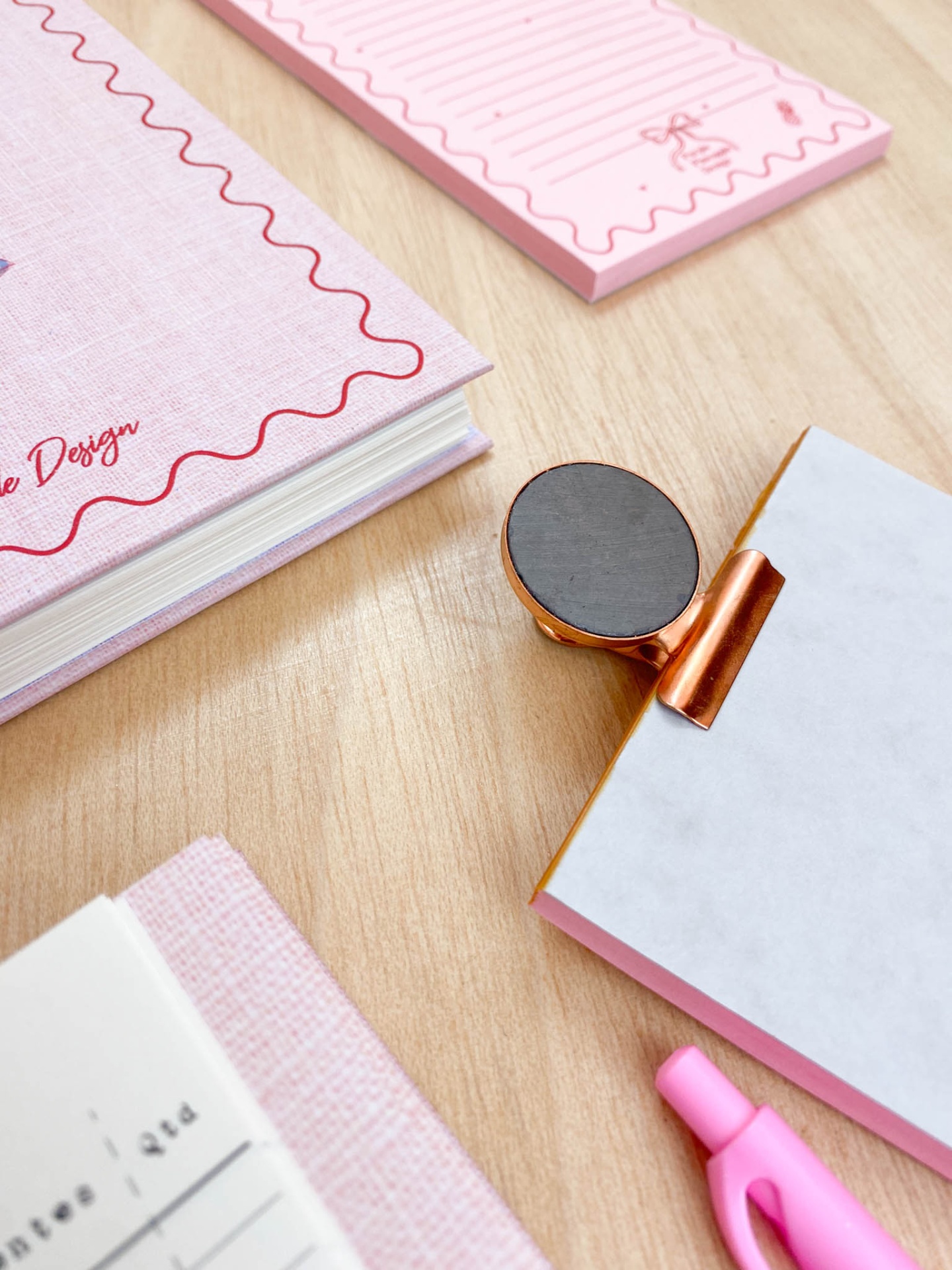Blocos de notas rosados com padrões ondulados, clipe metálico dourado com base magnética e caneta rosa sobre mesa de madeira