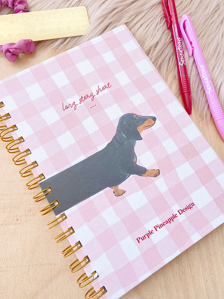 Caderno quadriculado com desenho de cão dachshund e texto em capa, canetas ao lado