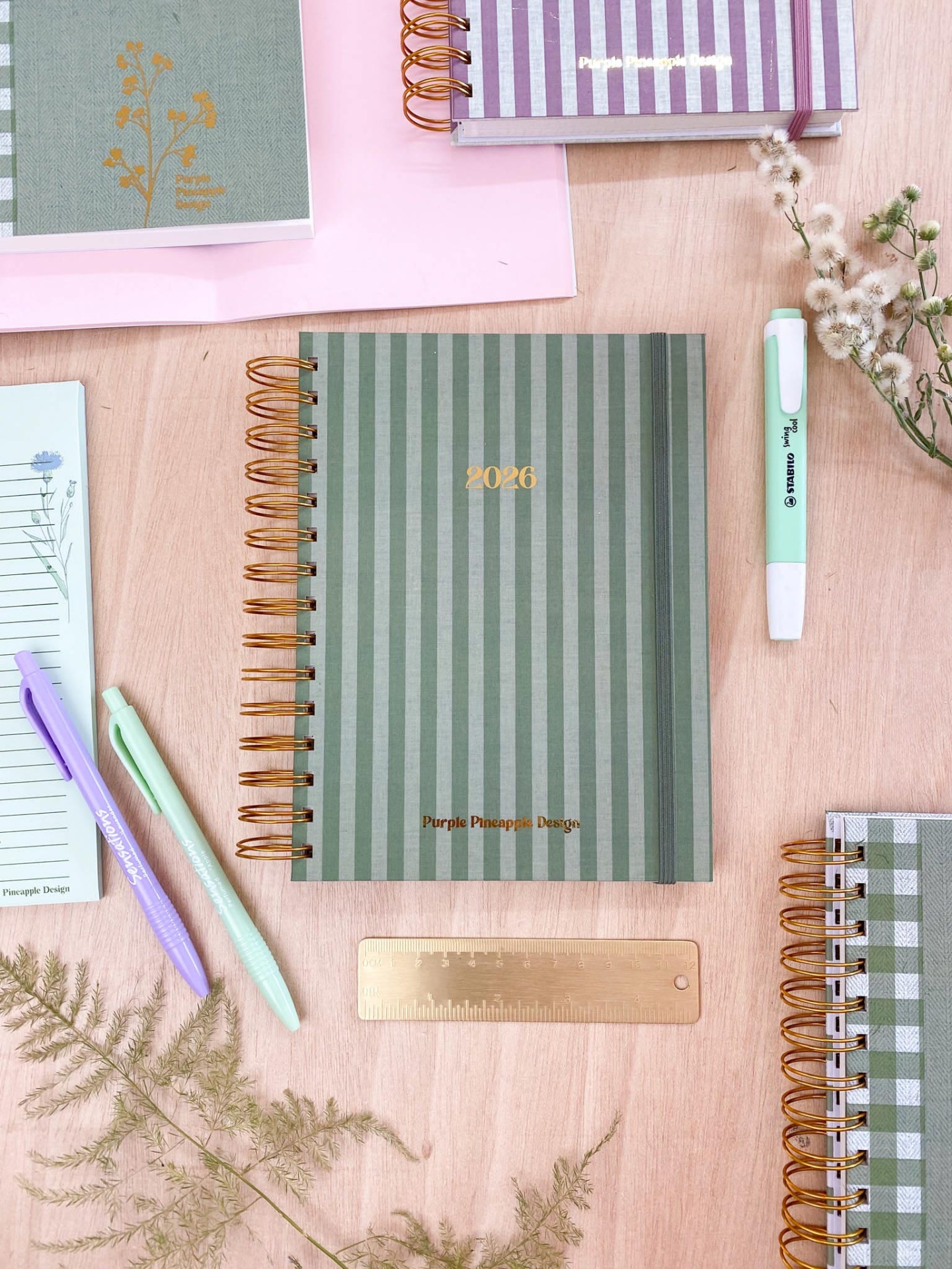 Caderno espiral riscas verde 2026 com canetas e plantas sobre mesa de madeira