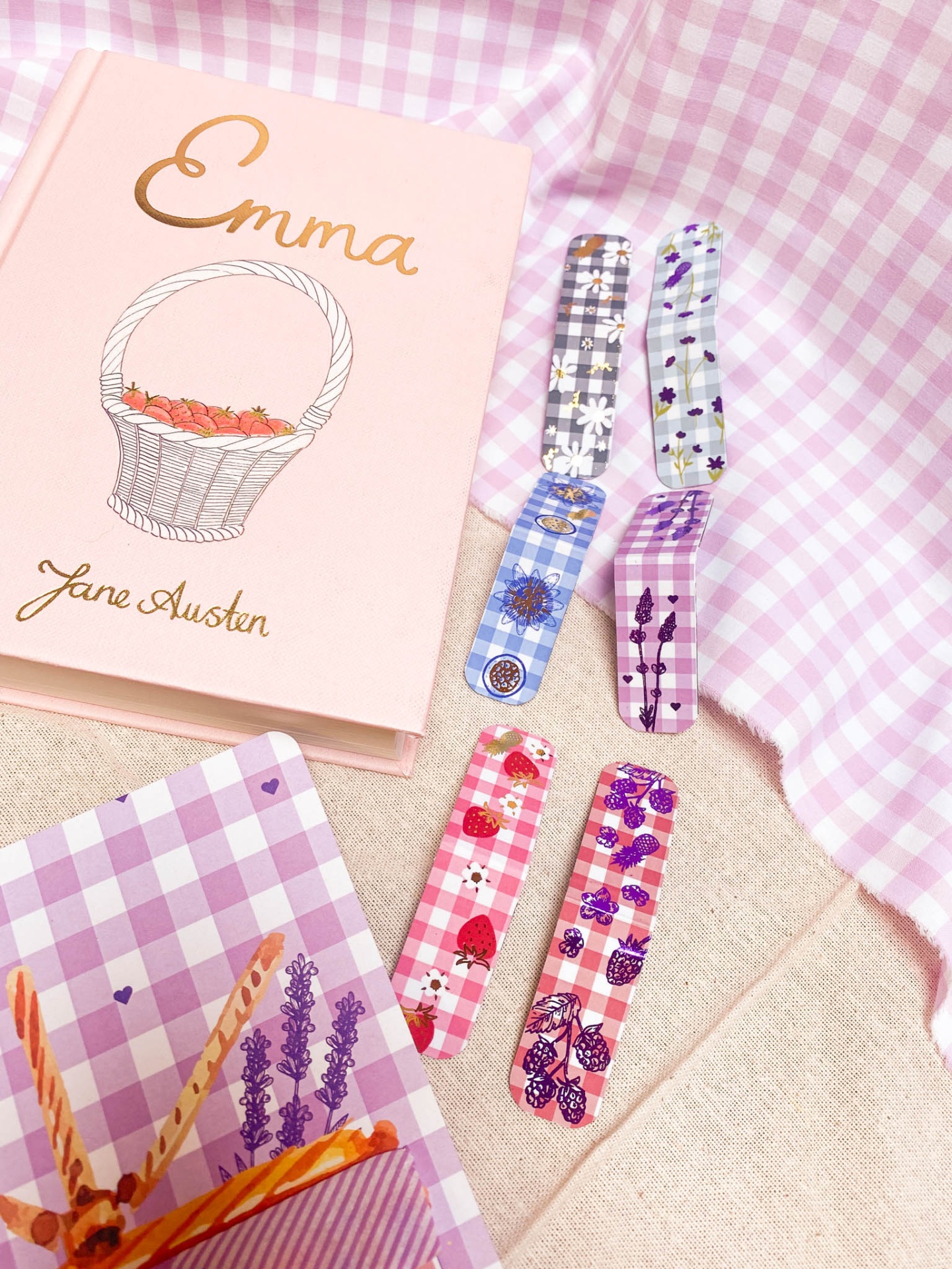 Livro rosa 'Emma' de Jane Austen ao lado de pregadores coloridos com padrões florais sobre tecido xadrez lilás.