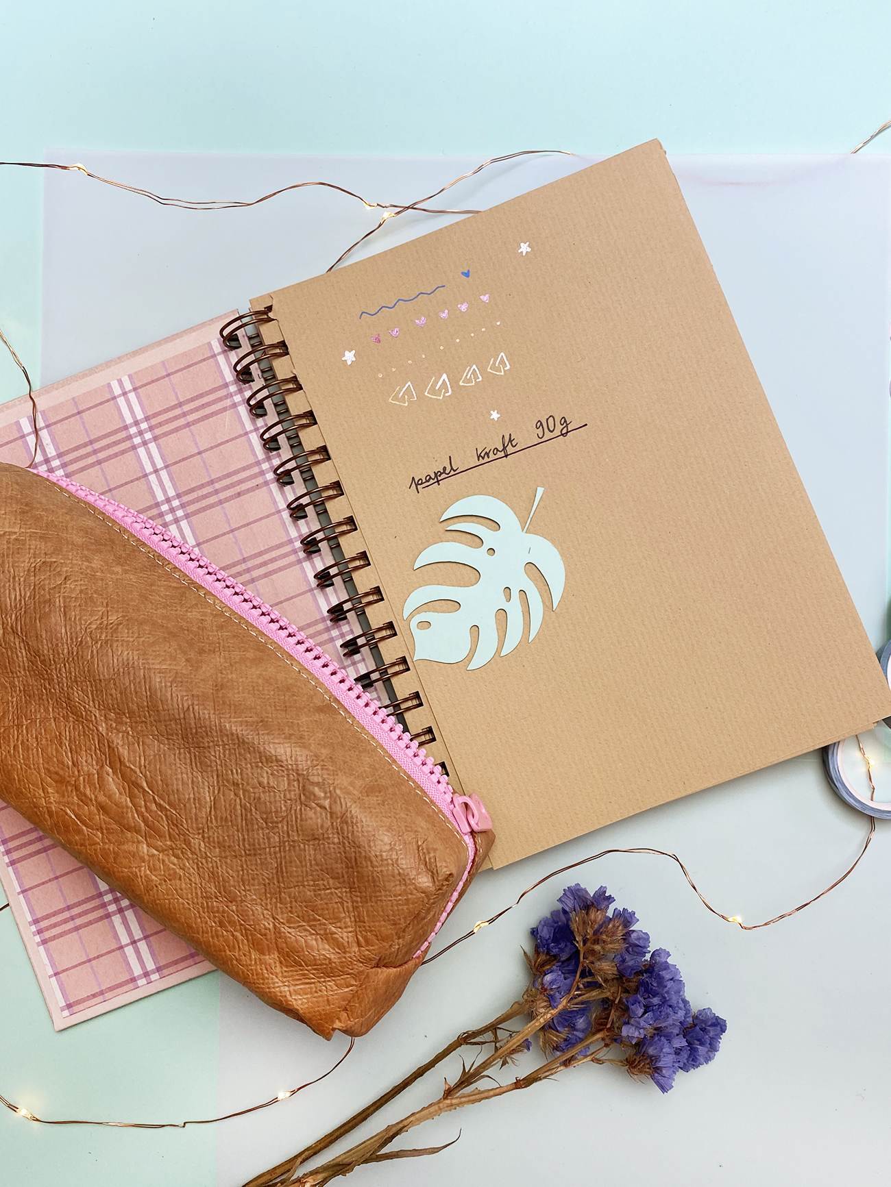 Dois cadernos em espiral, carteira castanha com fecho rosa, ramo de flores secas e luzes decorativas sobre superfície branca