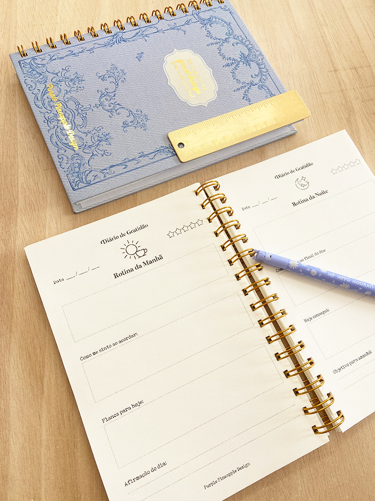 Caderno azul espiral aberto com rotinas diárias, régua dourada e caneta azul floral sobre mesa de madeira