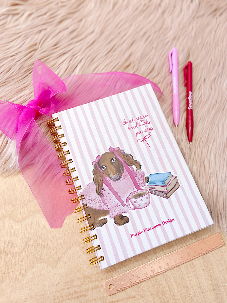 Como manter uma rotina criativa Caderno com capa de cão vestido de rosa, laço, canetas e régua sobre superfície clara