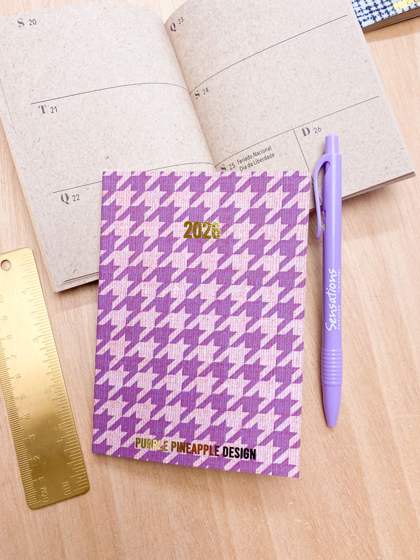 Agenda 2026 com capa lilás e rosa padrão pied-de-poule, caneta roxa e régua dourada sobre mesa de madeira