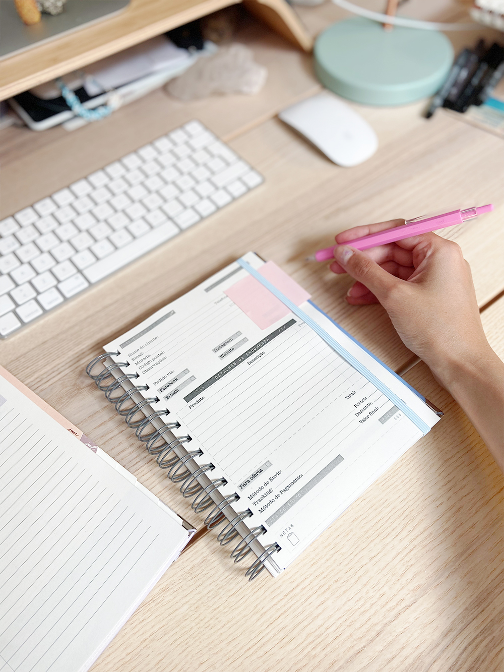 Caderno espiral aberto com formulário e caneta rosa numa secretária de madeira com teclado e rato branco