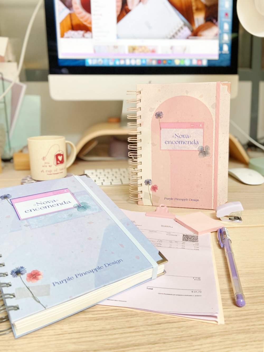 Dois cadernos espiralados rosa e azul com texto 'Nova encomenda' numa mesa de madeira com computador ao fundo