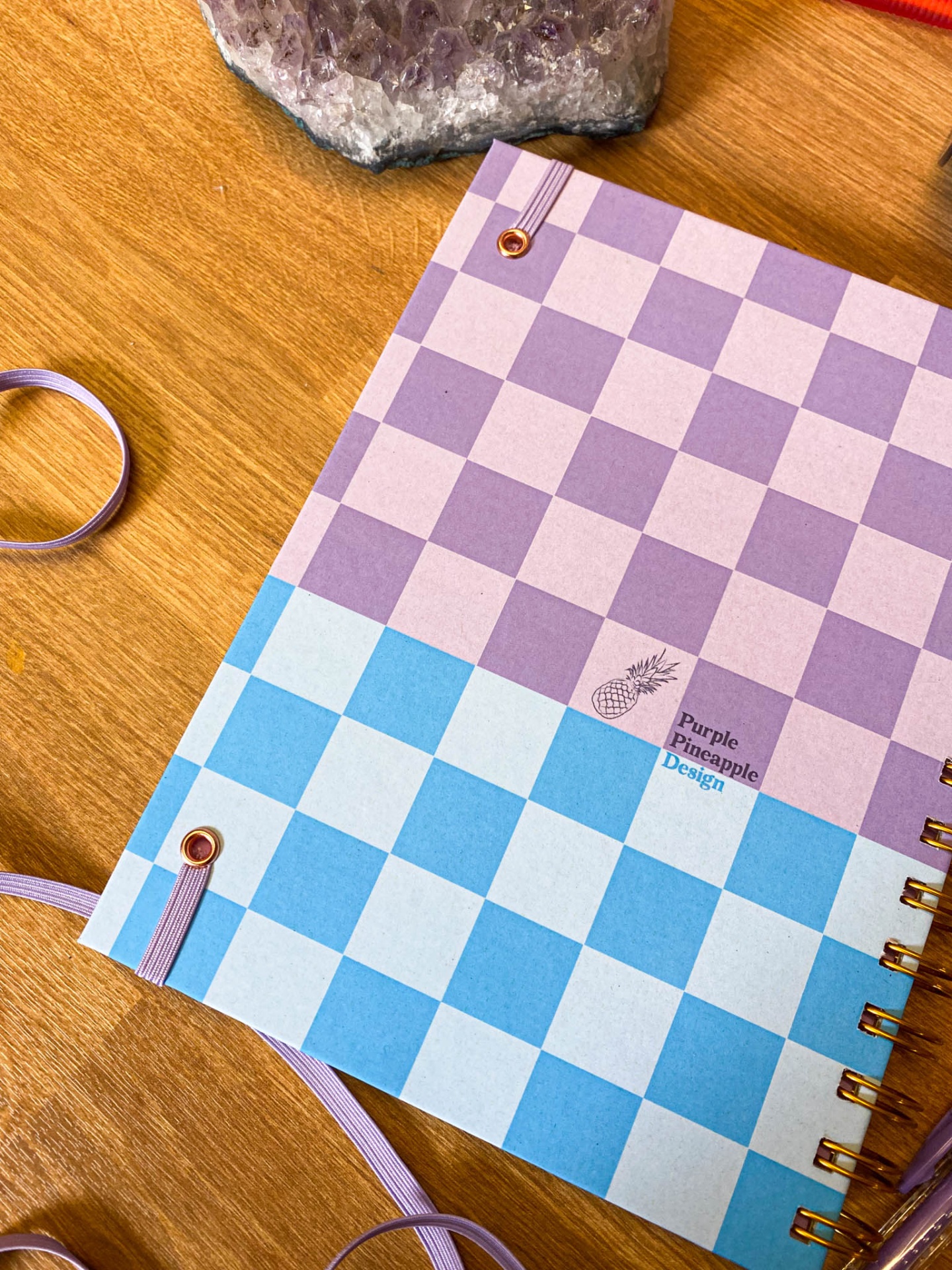 Caderno quadriculado lilás, rosa e azul claro com texto Purple Pineapple Design sobre mesa de madeira