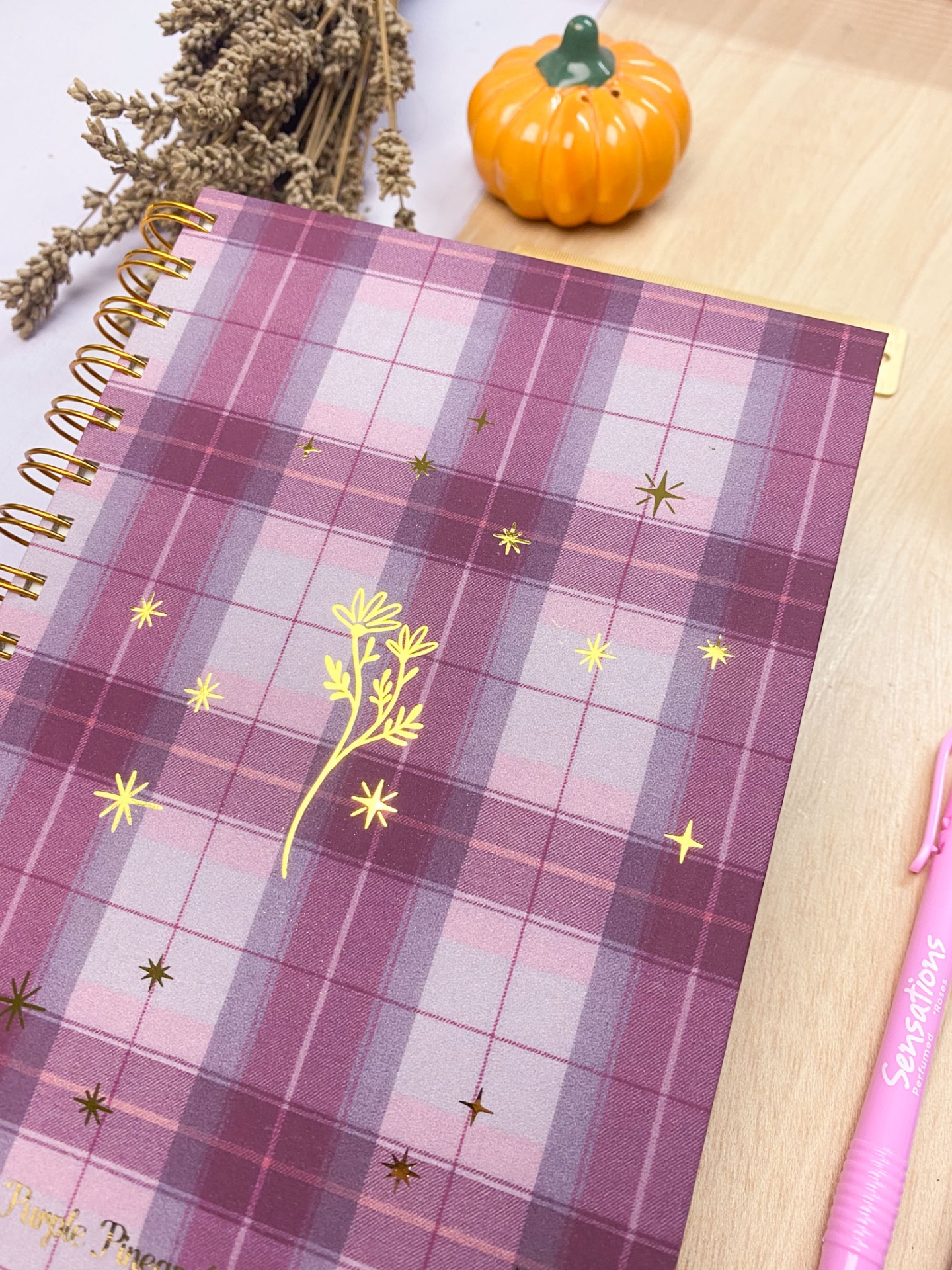 Caderno xadrez roxo com detalhes dourados e caneta rosa sobre mesa de madeira