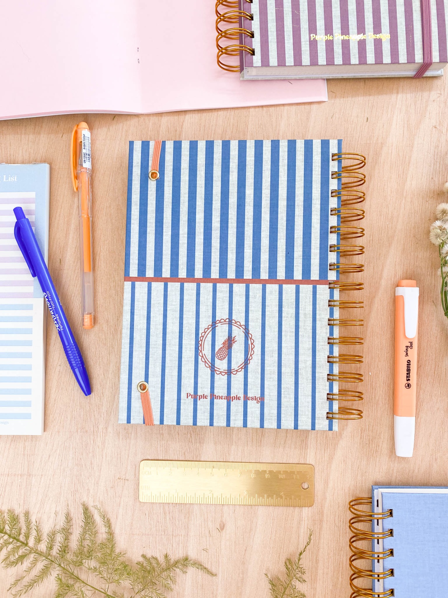 Cadernos às riscas azul e branco com decoração castanha, canetas coloridas, régua dourada e planta sobre mesa de madeira