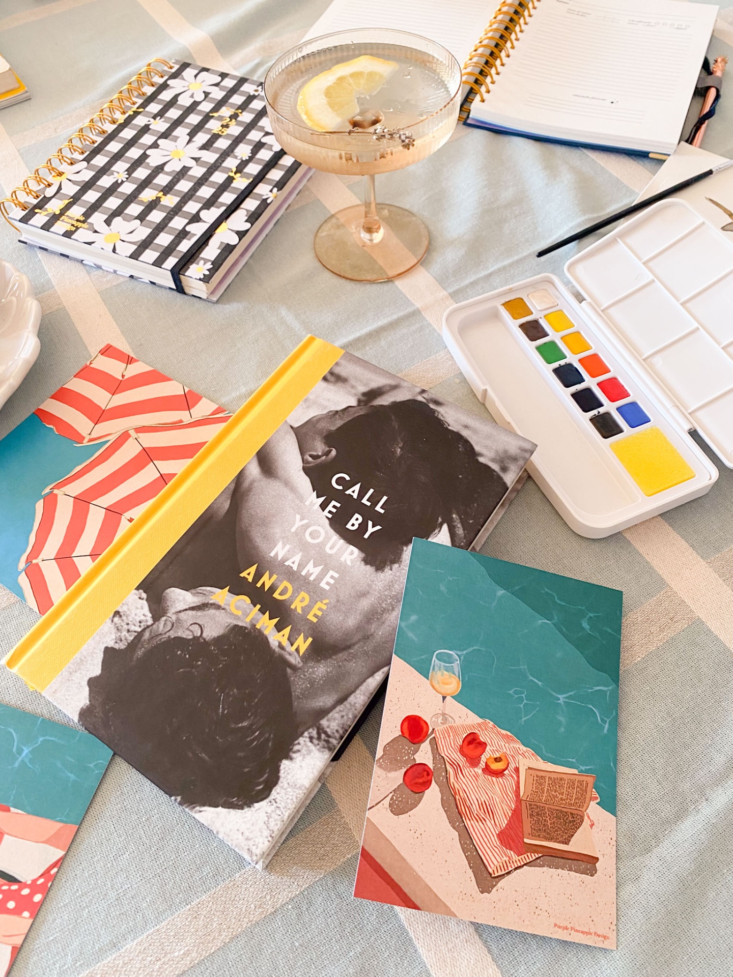 Mesa com livro, cadernos, copo com bebida, paleta de tintas, caneta e cartão postal