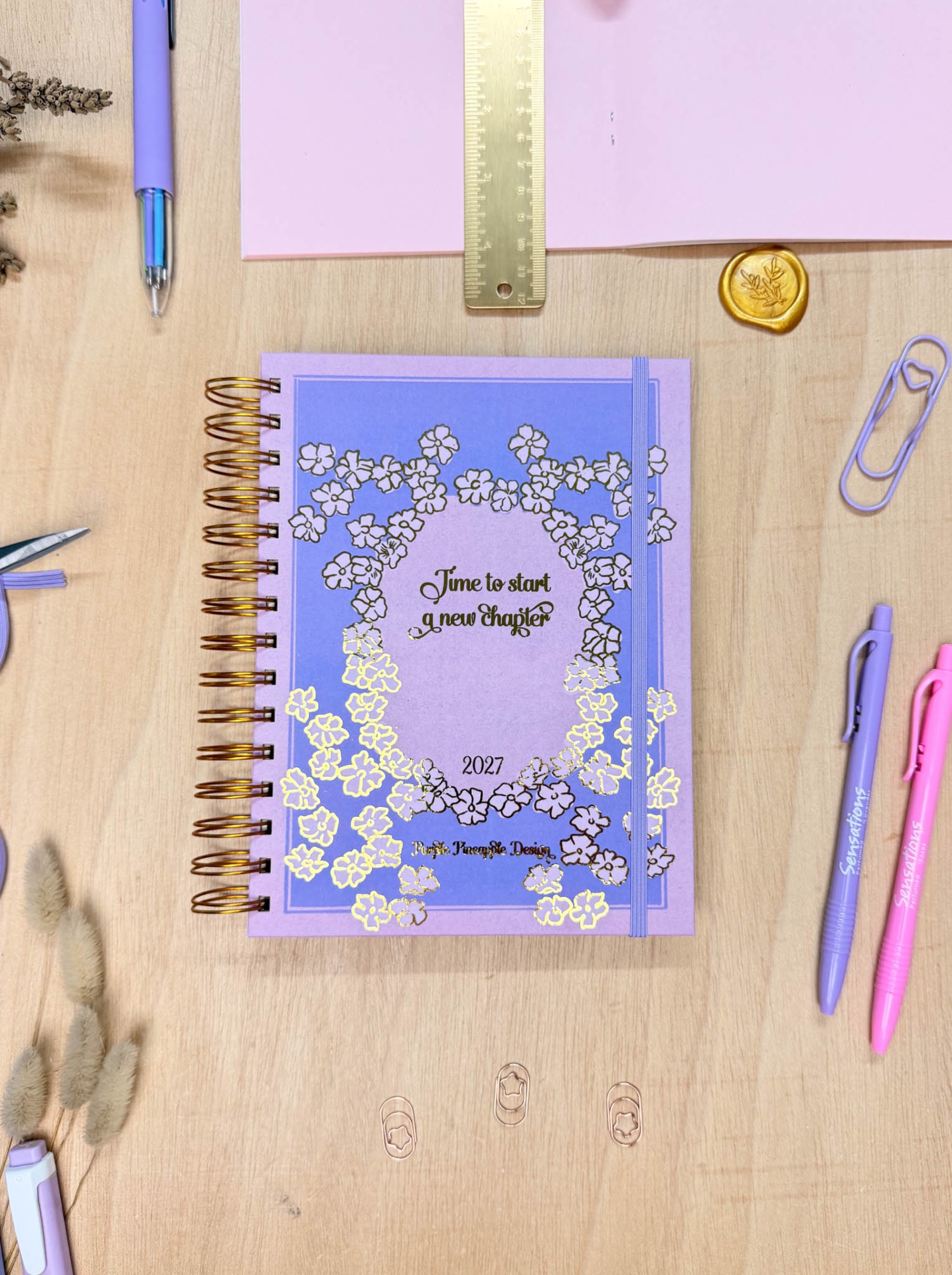 Agenda anual roxa espiralada com flores douradas na mesa ao lado de canetas e clips