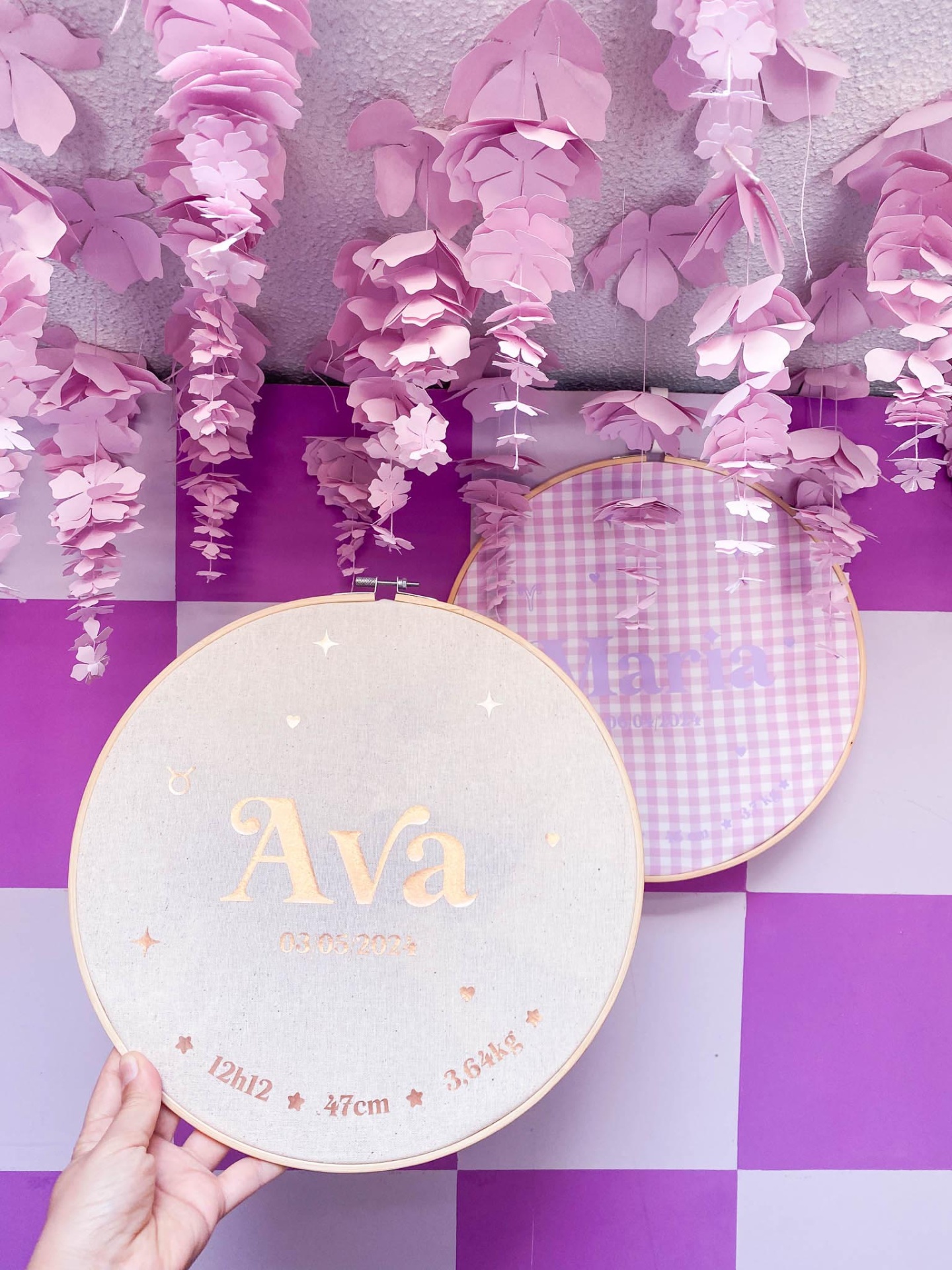 Dois aros de bordado com nome e padrão xadrez, decoração floral cor-de-rosa no teto e parede quadriculada roxa
