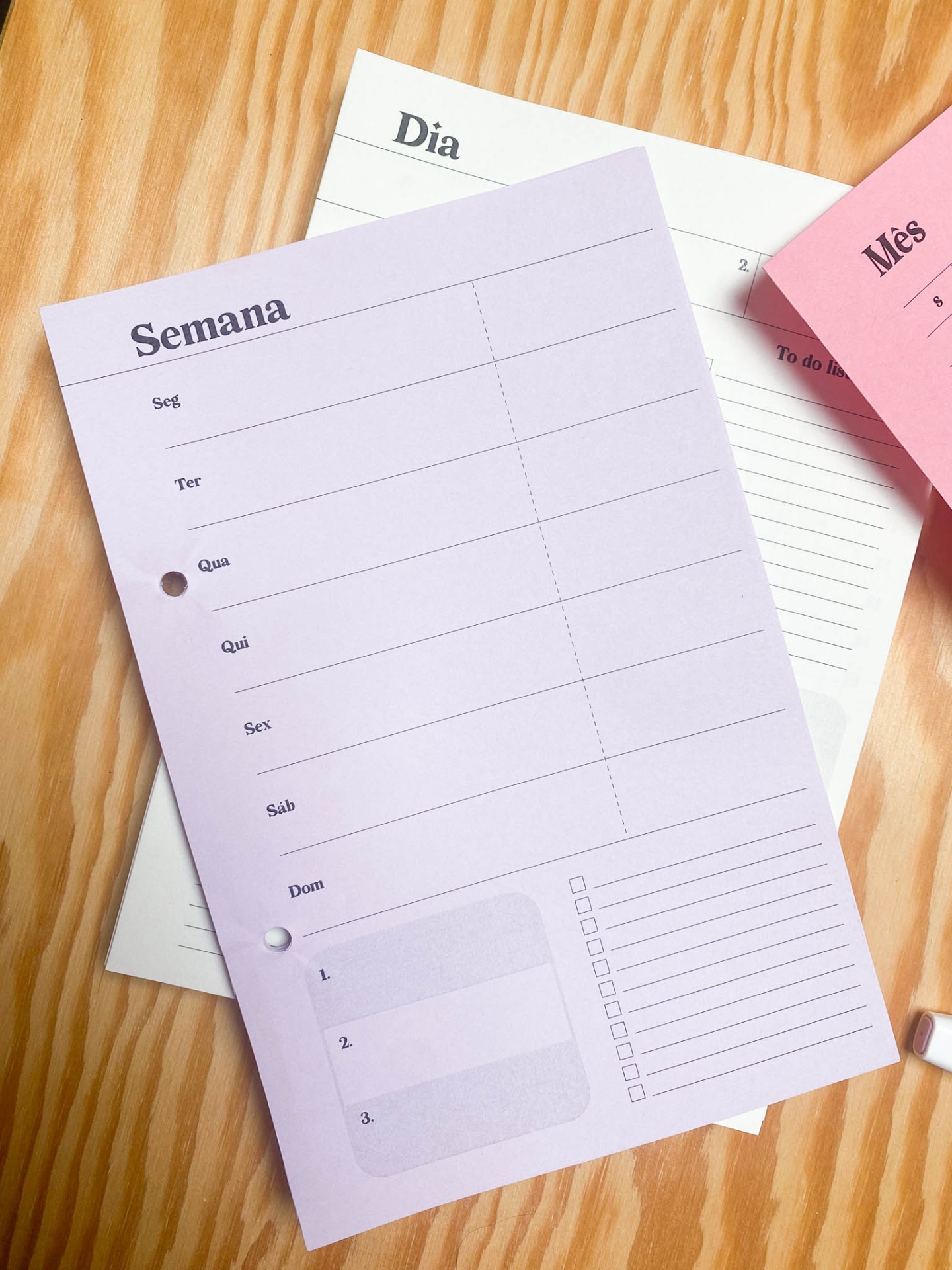 Folhas de papelaria para planeamento semanal, diária e mensal em tons de rosa e lilás sobre mesa de madeira