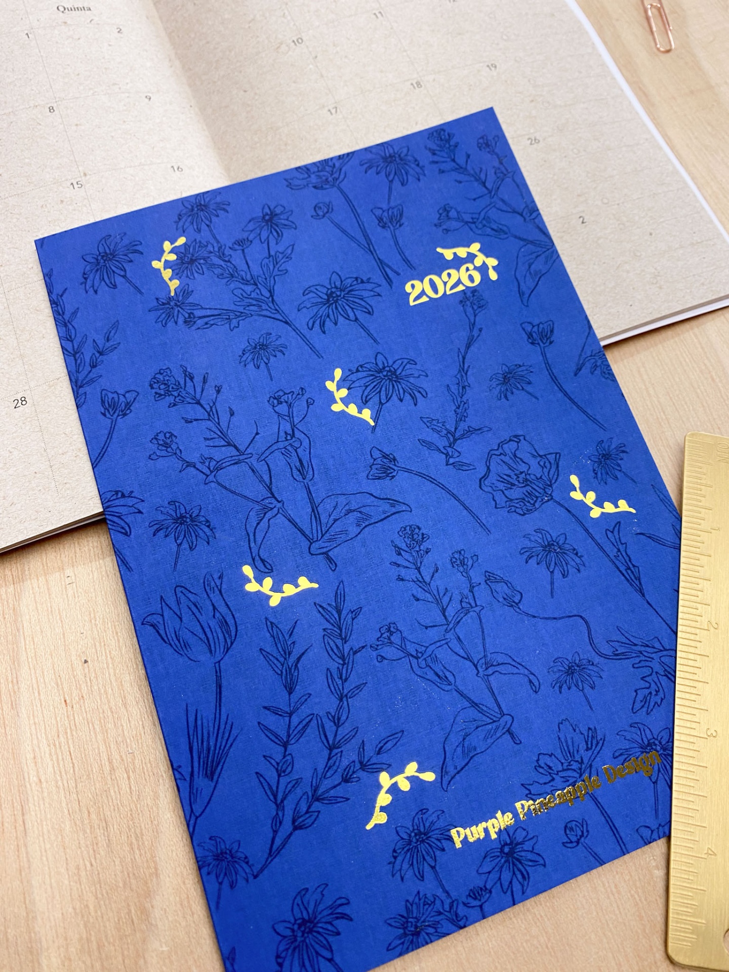 Caderno azul com estampado floral e texto amarelo '2026' e 'Purple Planner Company' sobre mesa de madeira
