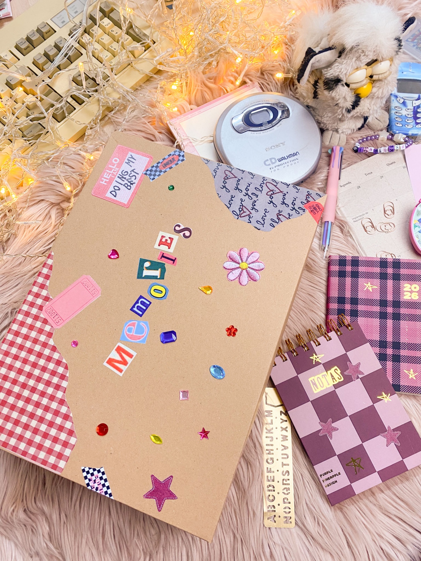 Caderno decorado com colagens, CD Walkman, peluche, canetas e calendários numa superfície peluda rosa com luzes.