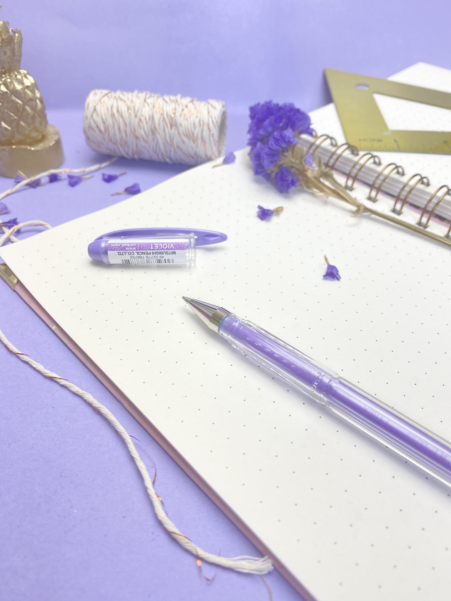Caderno pontilhado com caneta roxa, marcador NOTICE, fio decorativo, flores secas e régua dourada sobre superfície lilás