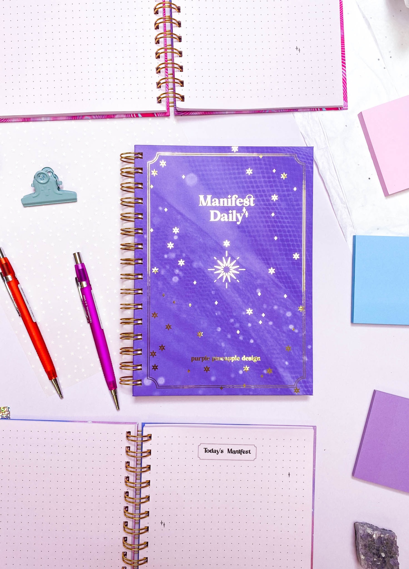 Caderno roxo Manifest Daily com estrelas douradas em fundo branco com canetas e blocos de notas coloridos
