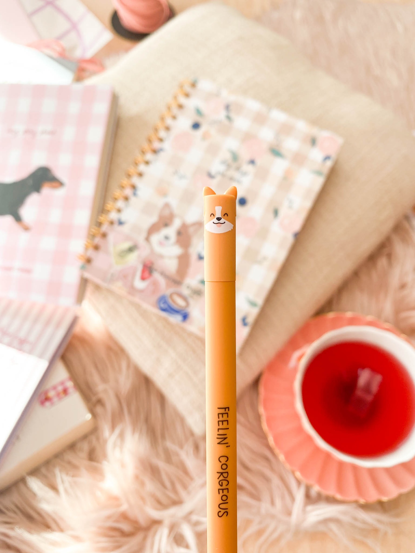 Caneta laranja com tampa em forma de cabeça de cão e texto FEELIN' GORGEOUS, com fundo desfocado de caderno xadrez e chá em xícara rosa.