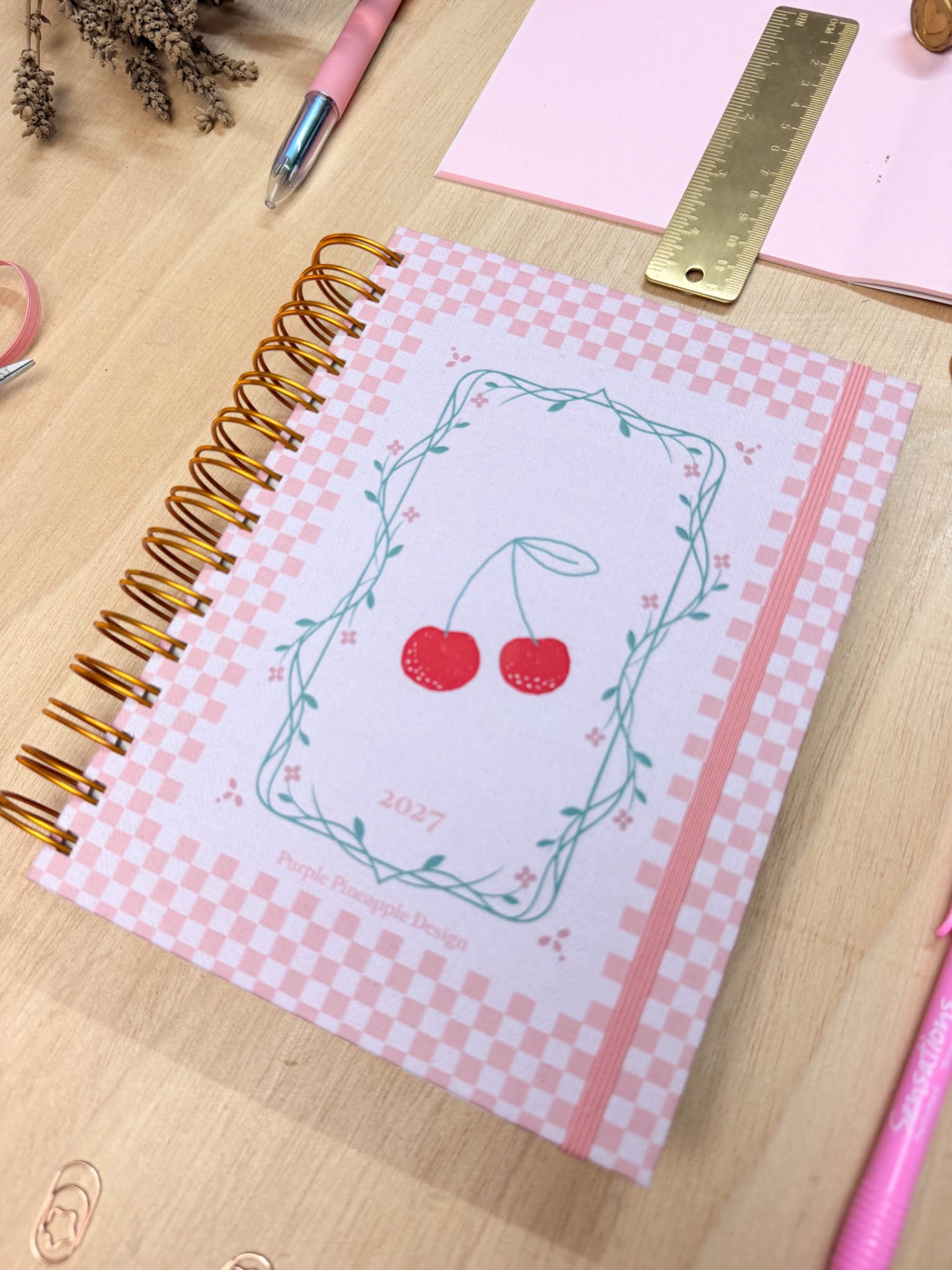 Caderno espiral com capa xadrez rosa e branco com cerejas e texto 2027 sobre mesa com régua e canetas.