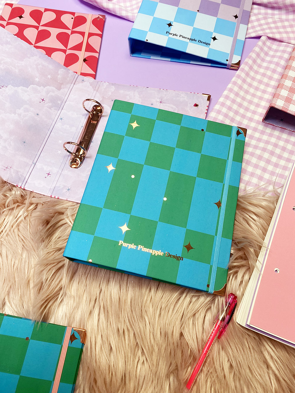 Cadernos tipo fichário com capas coloridas em padrões quadriculados e de coroa, sobre superfície peluda e fundo lilás.