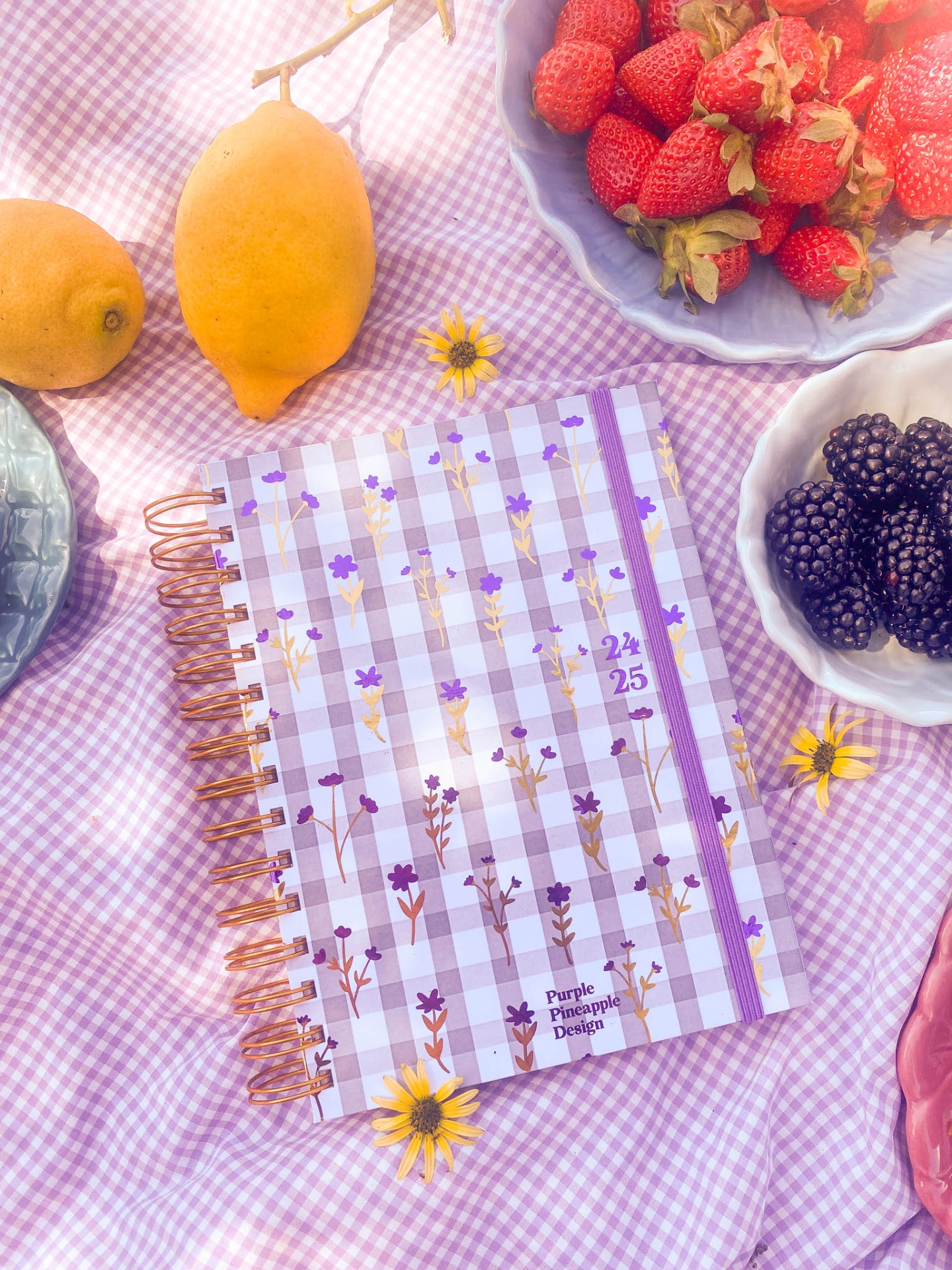 Agenda espiral com capa xadrez lilás e flores sobre tecido cor-de-rosa com frutas e flores ao redor.